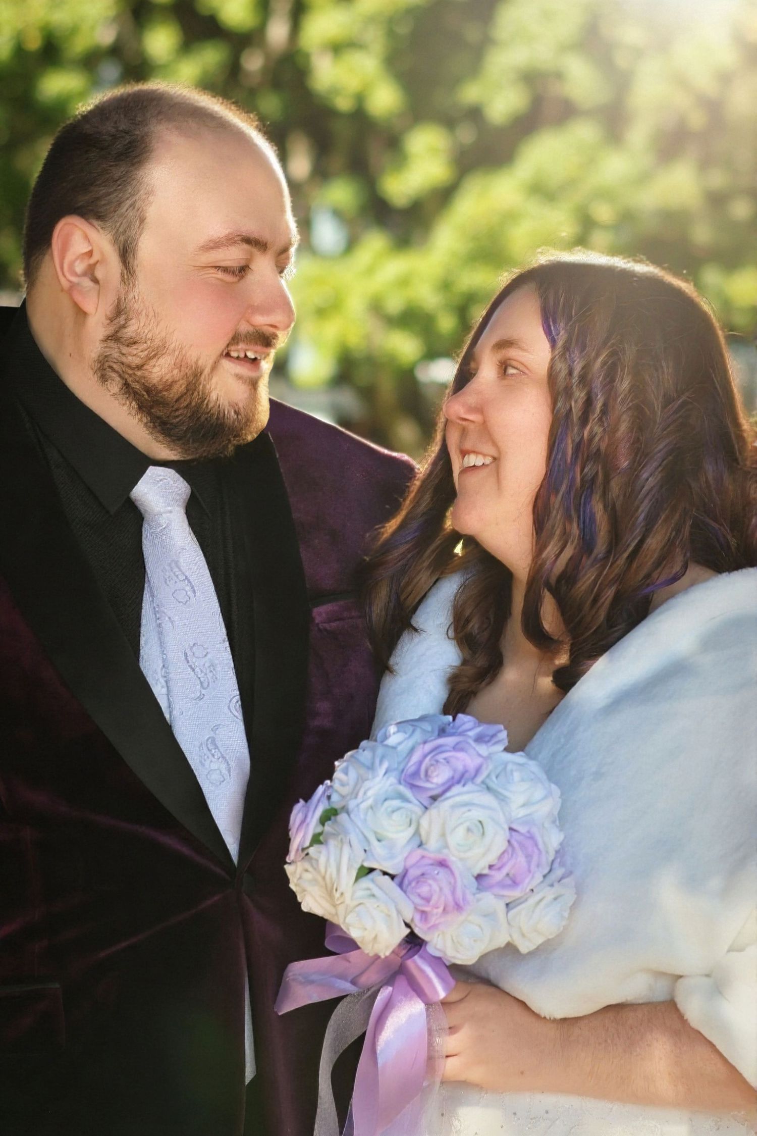 Smiling bride and groom in sunlit garden, groom in burgundy velvet suit, bride holding purple and white bouquet.