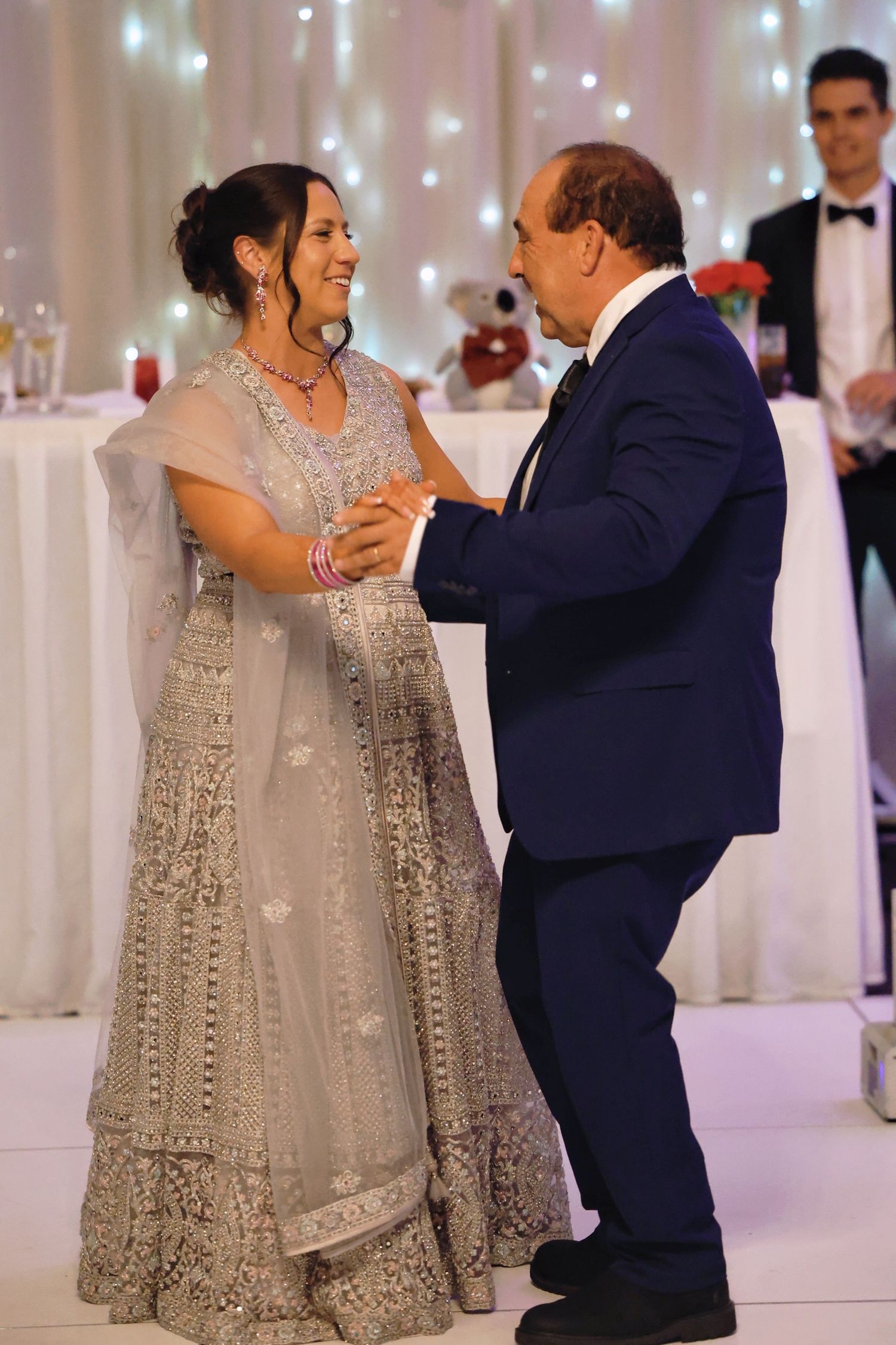 Bride in silver lehenga dancing with man in navy suit at elegant wedding reception with fairy lights.