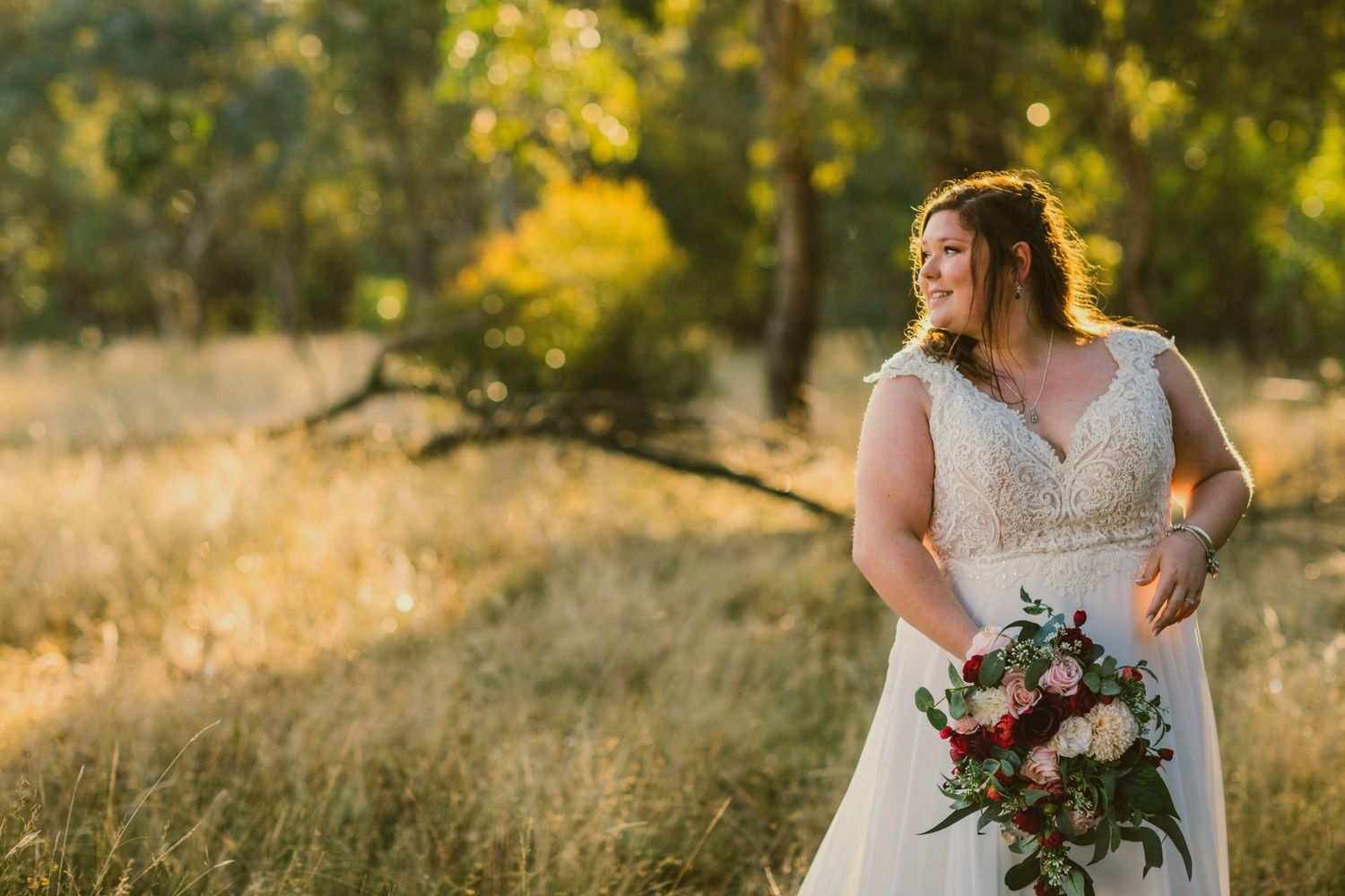 Radiant bride in lace wedding gown holds bouquet of red and pink roses in a sunlit golden field.