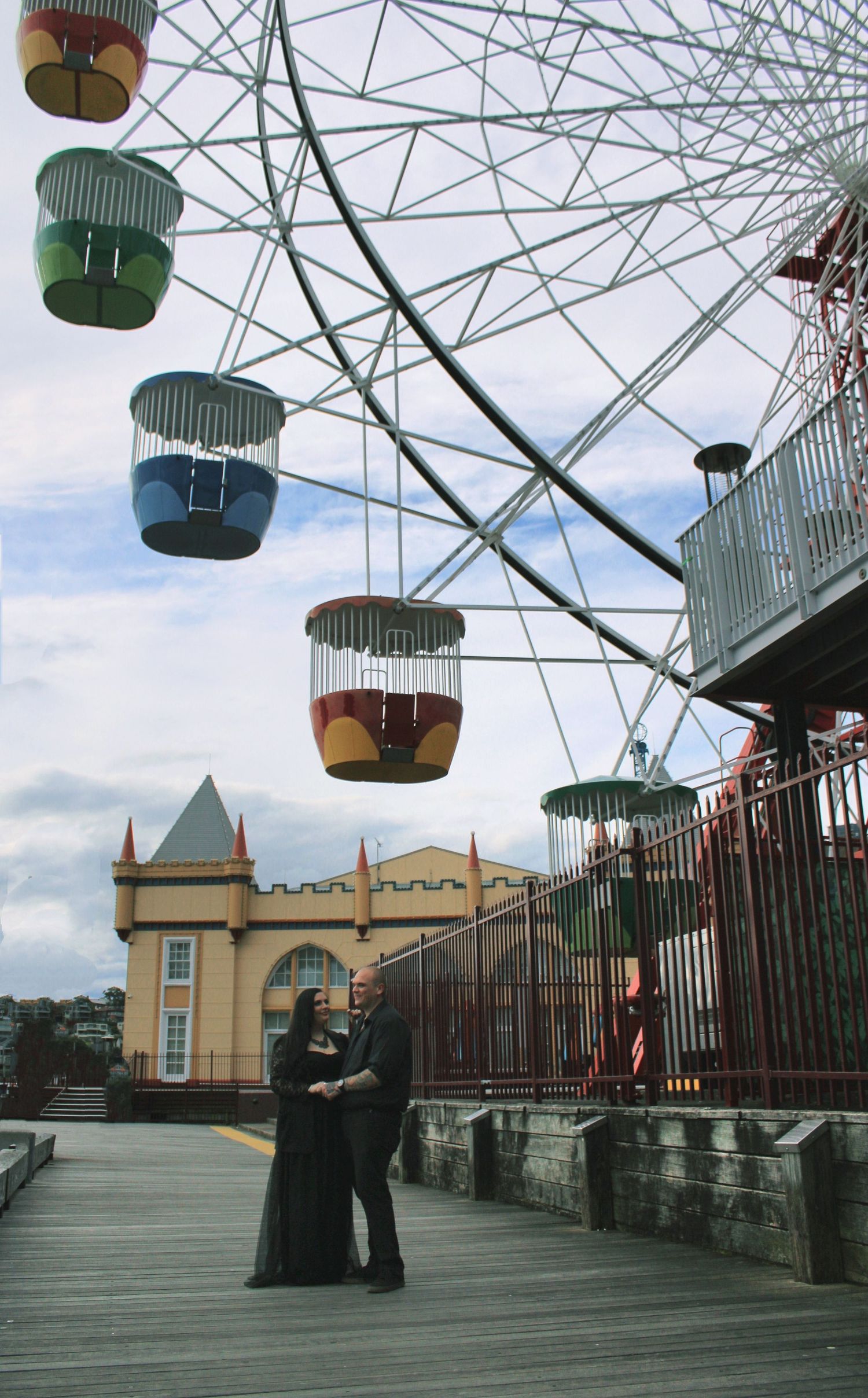 Couple embracing beneath colorful Ferris wheel gondolas at amusement park boardwalk.
