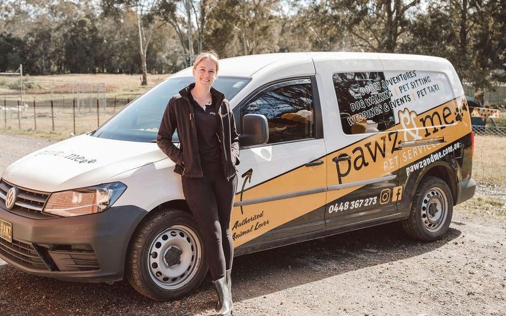 Woman standing beside a Pawz & Me mobile pet grooming van parked outdoors in a rural Australian setting.