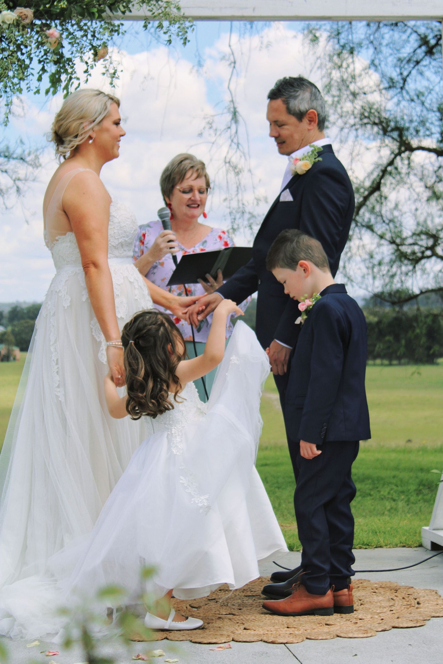 Bride and groom exchange rings at outdoor ceremony altar with two children and officiant present.