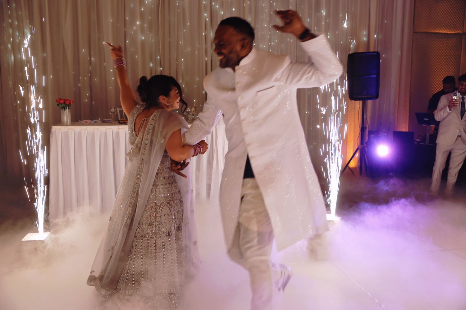 Couple dancing at wedding reception with sparklers and fog machine effects in elegant ballroom.