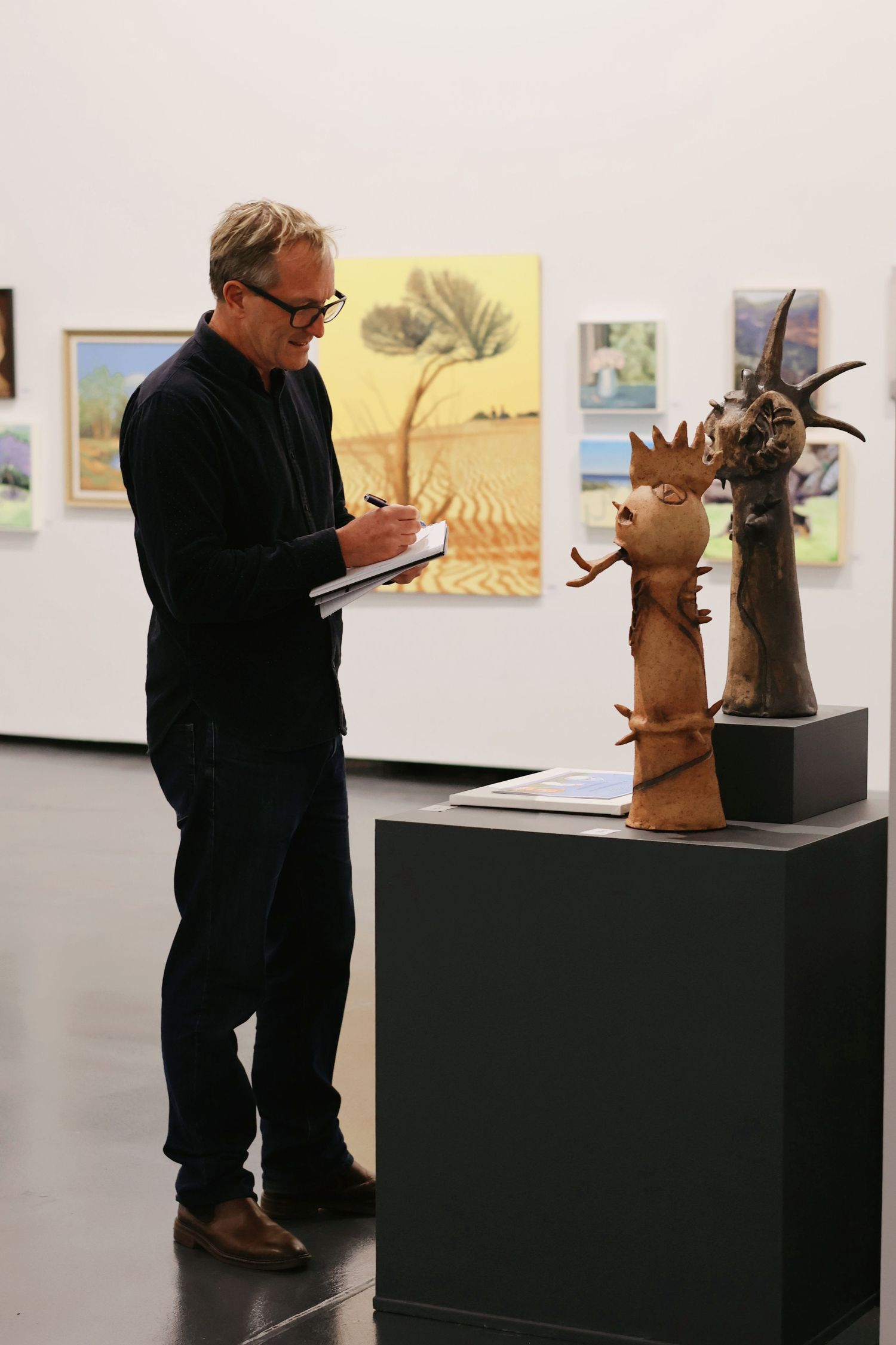 Man taking notes while examining abstract ceramic sculptures on a pedestal in a contemporary art gallery.