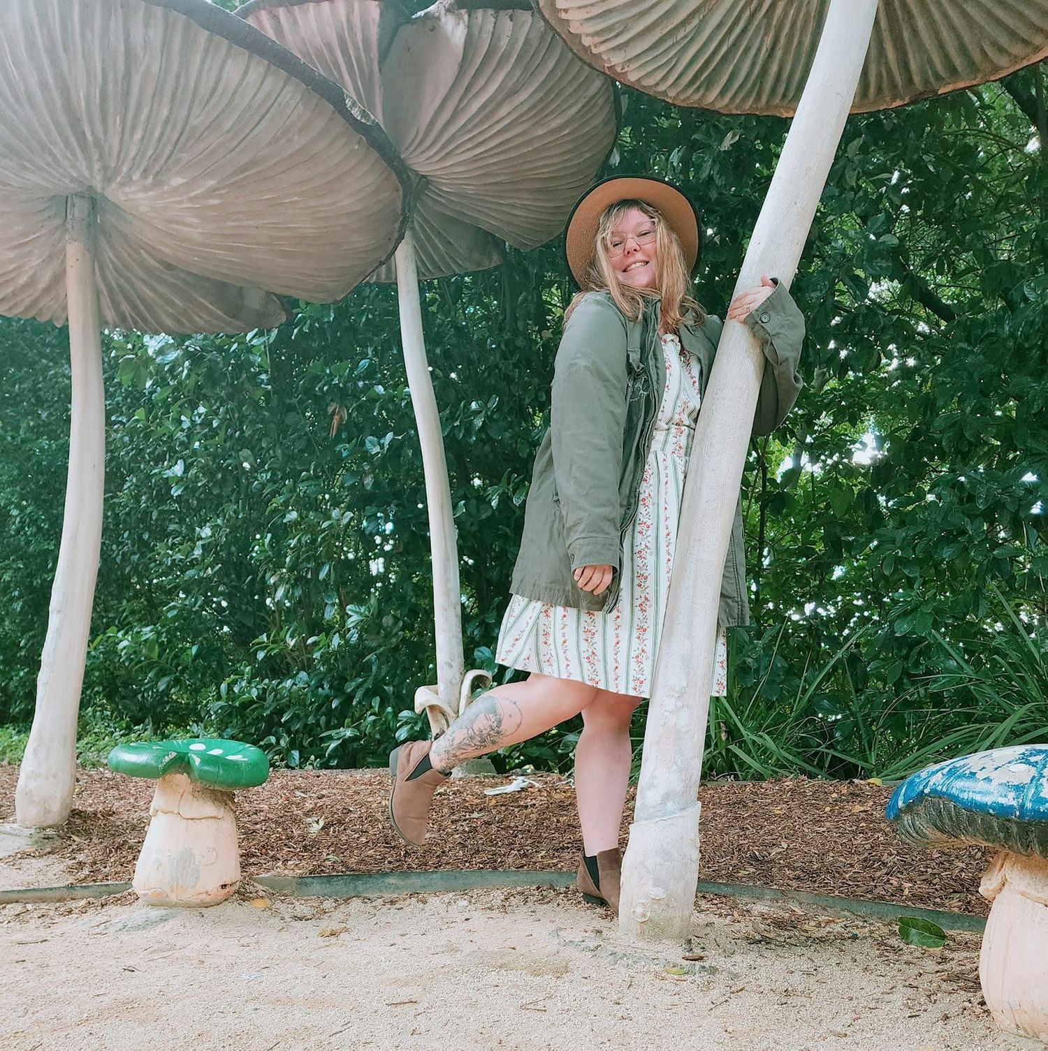 Woman in hat and dress poses among giant mushroom sculptures in an outdoor garden setting.