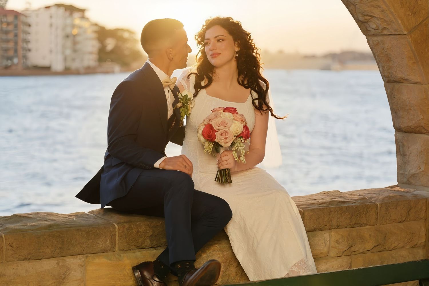 Bride and groom sitting on stone wall by waterfront at golden sunset holding wedding bouquet.
