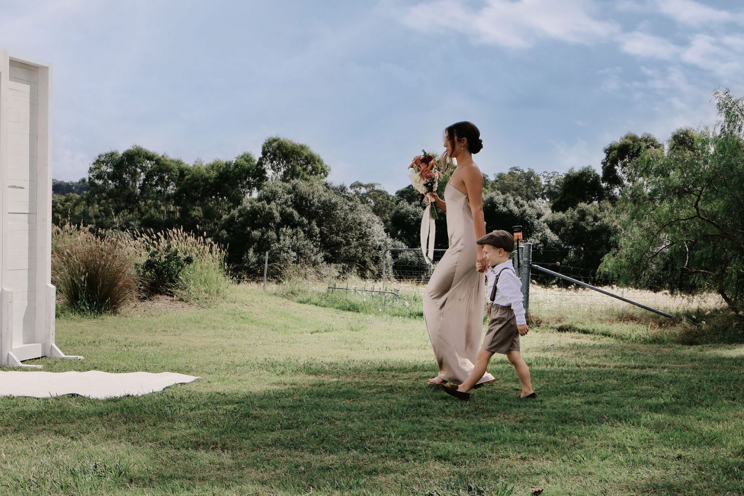 Woman in a beige dress walking with a young boy in suspenders across a green lawn at an outdoor wedding venue.