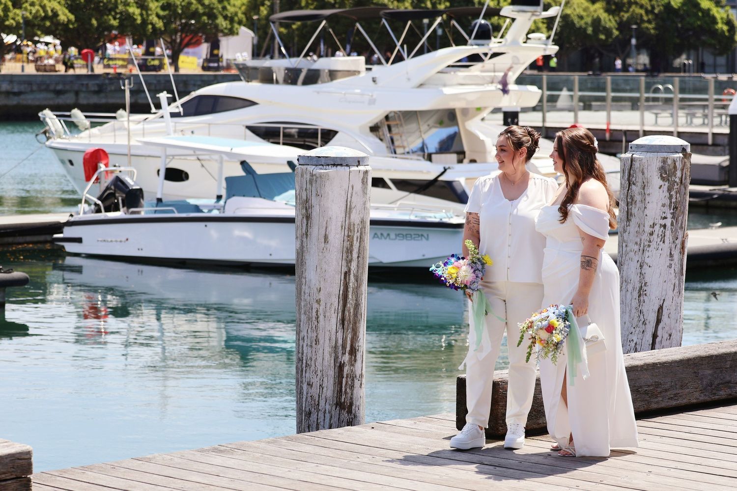 Two brides in white holding floral bouquets standing on a marina dock with luxury yachts in the background.