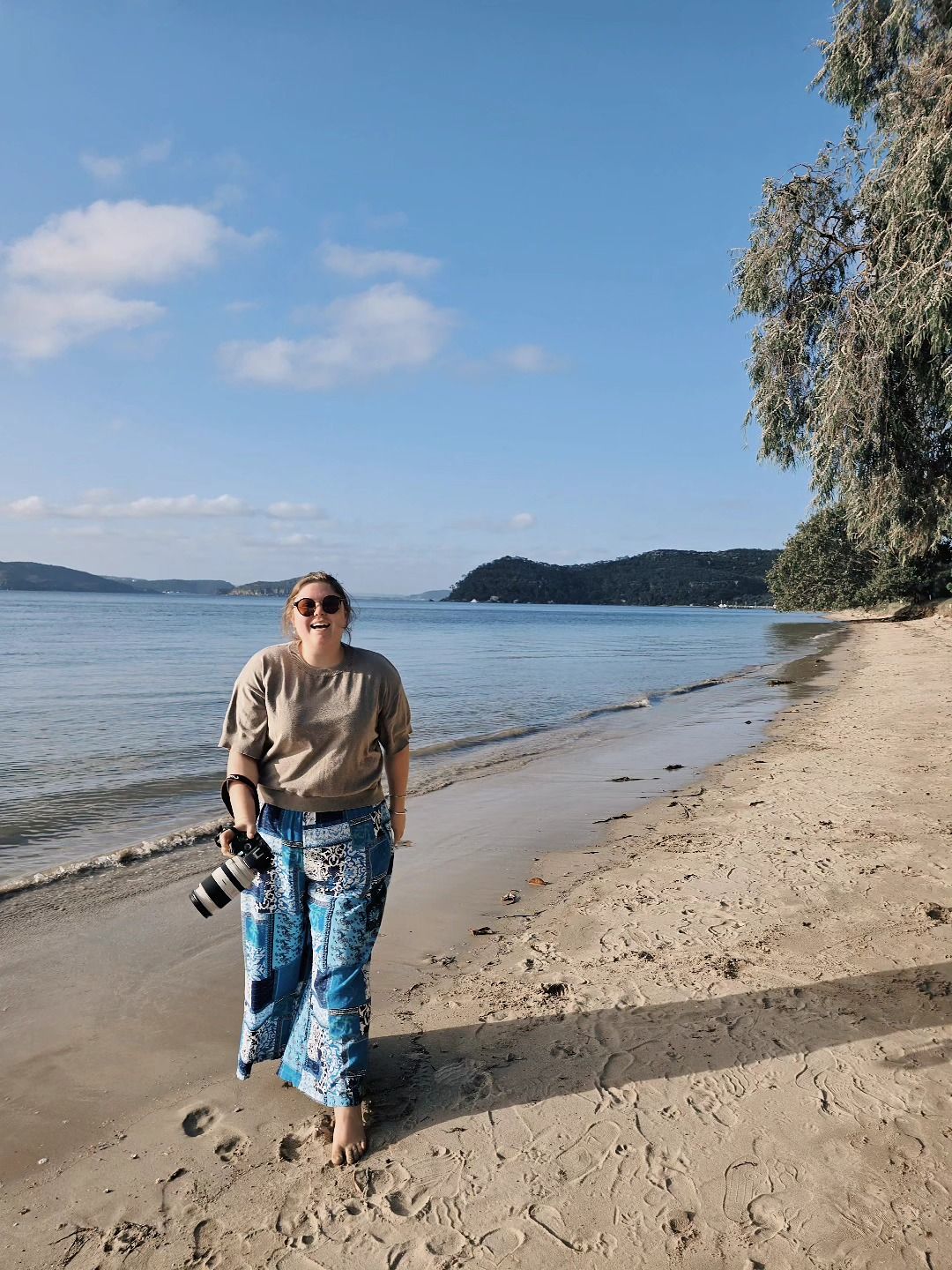 Woman walking barefoot on a sandy beach holding a camera, with calm blue water and trees behind her.