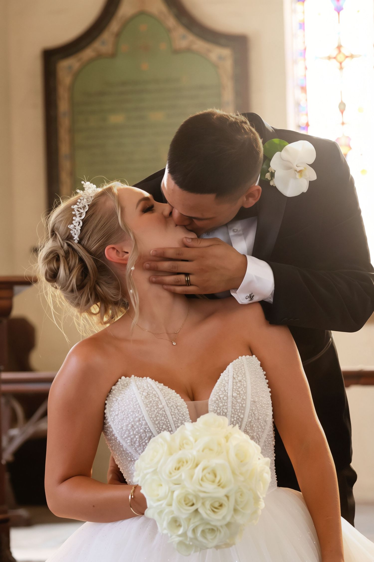 Groom in black tuxedo kisses bride wearing pearl-beaded gown and tiara inside a church.