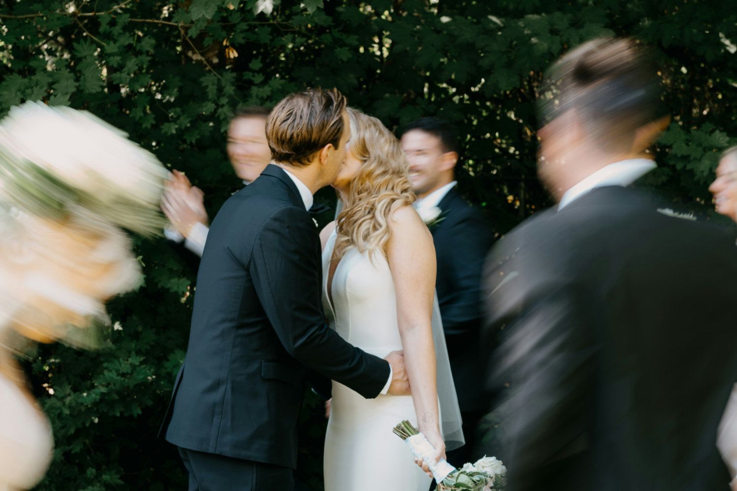 A bride and groom share a kiss surrounded by blurred guests in a lush garden setting.