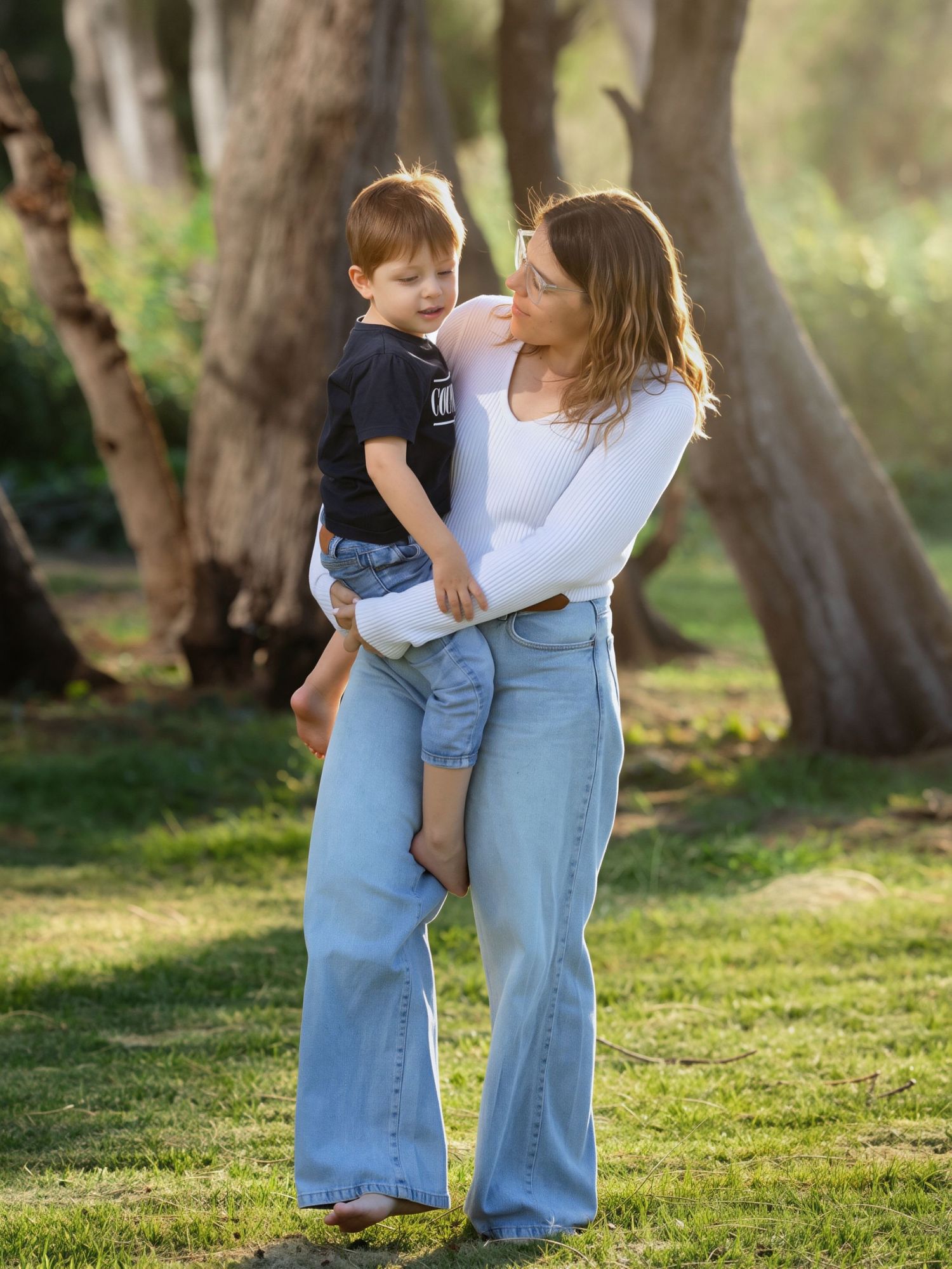 A mother in a white top and wide-leg jeans tenderly holds her young son in a sunlit park.