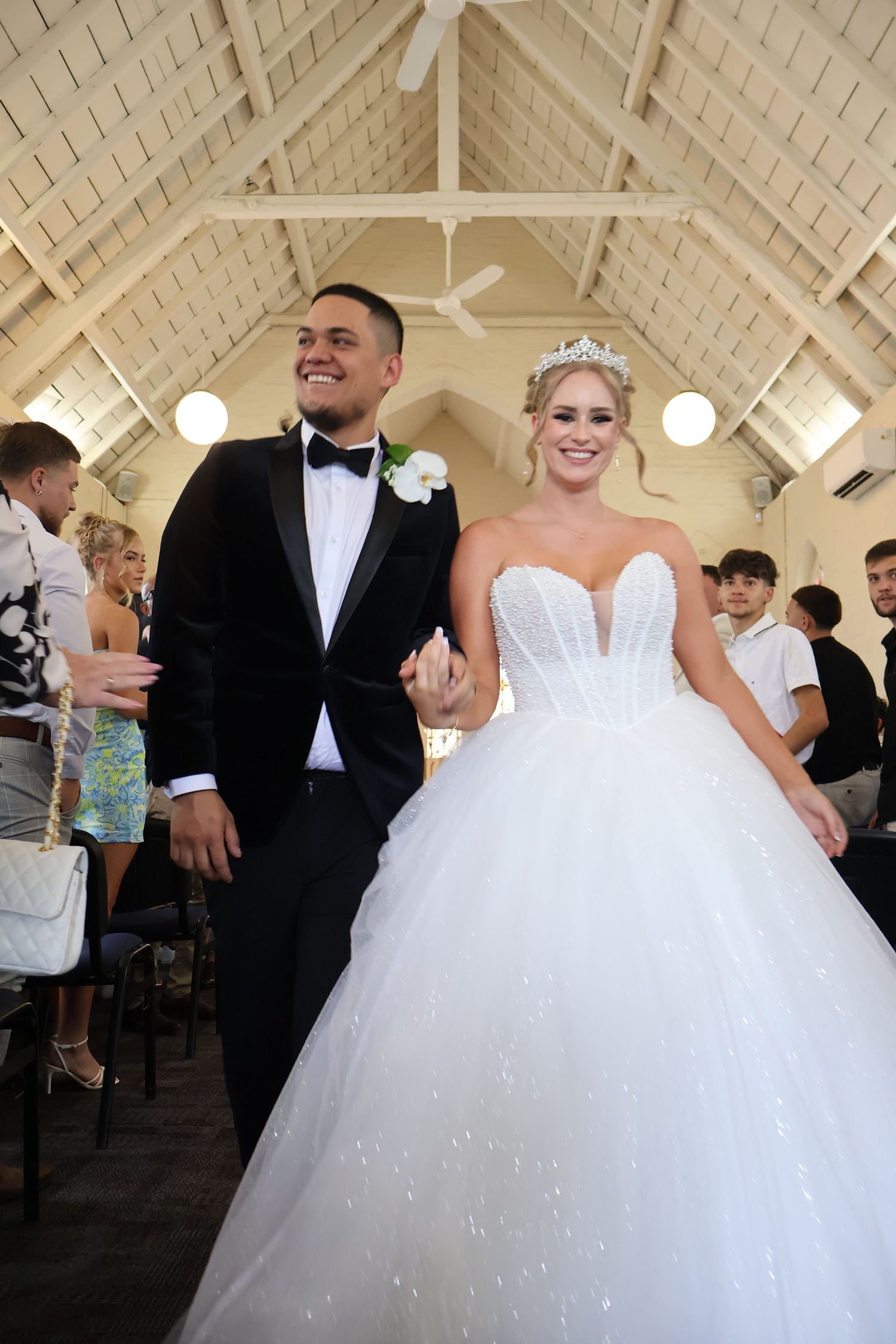 Smiling newlyweds walk down church aisle, bride in sparkling ball gown with tiara, groom in black tuxedo.