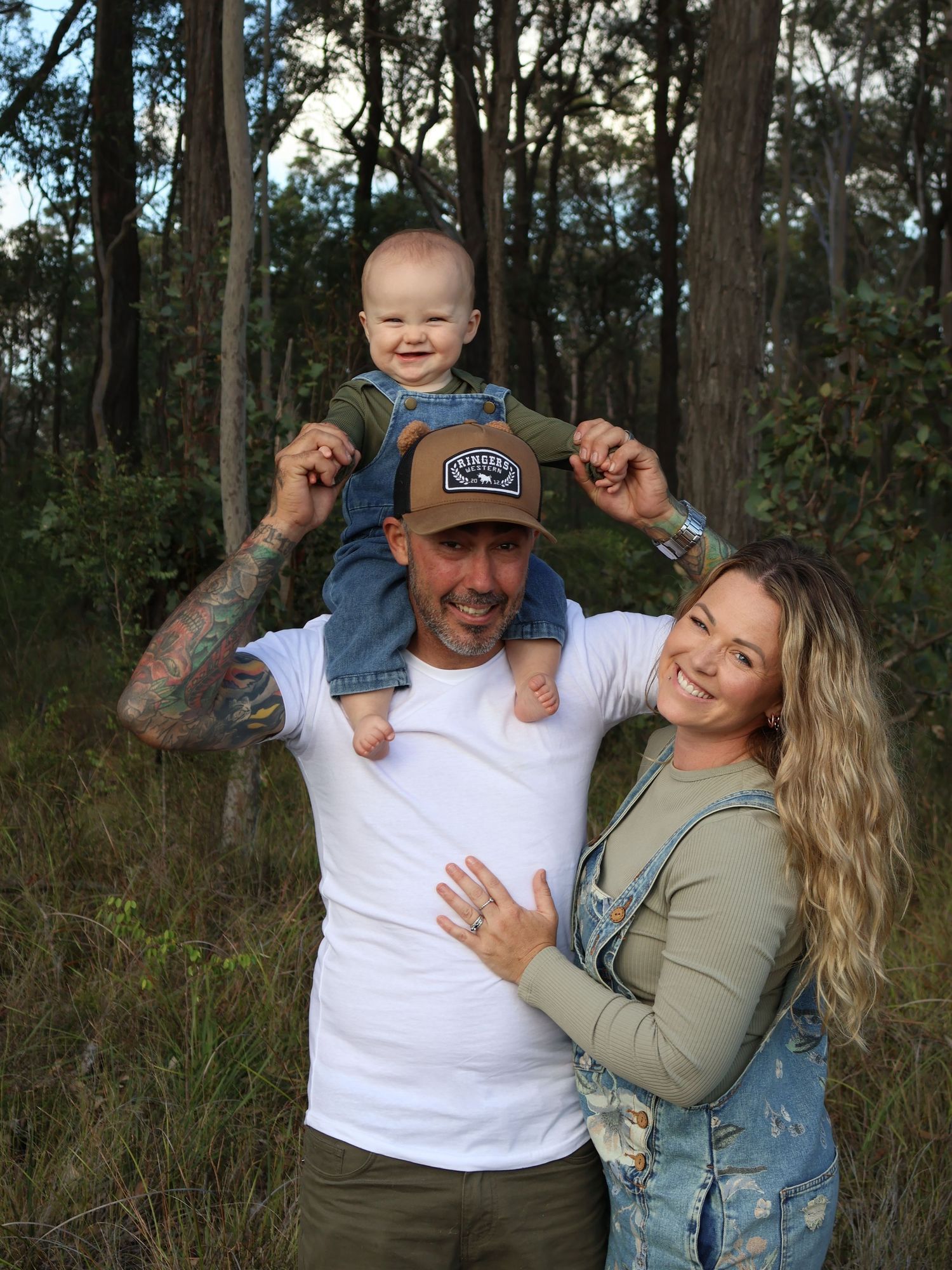 Happy family with tattooed dad holding baby on shoulders, mom embracing them in a forest setting.