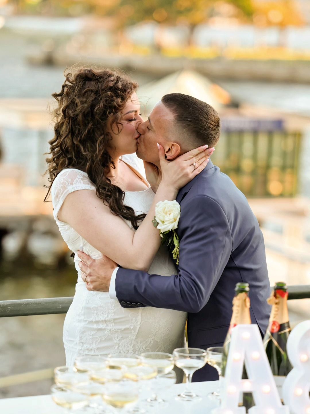 Bride and groom share a romantic kiss at an outdoor waterfront wedding reception with champagne glasses.