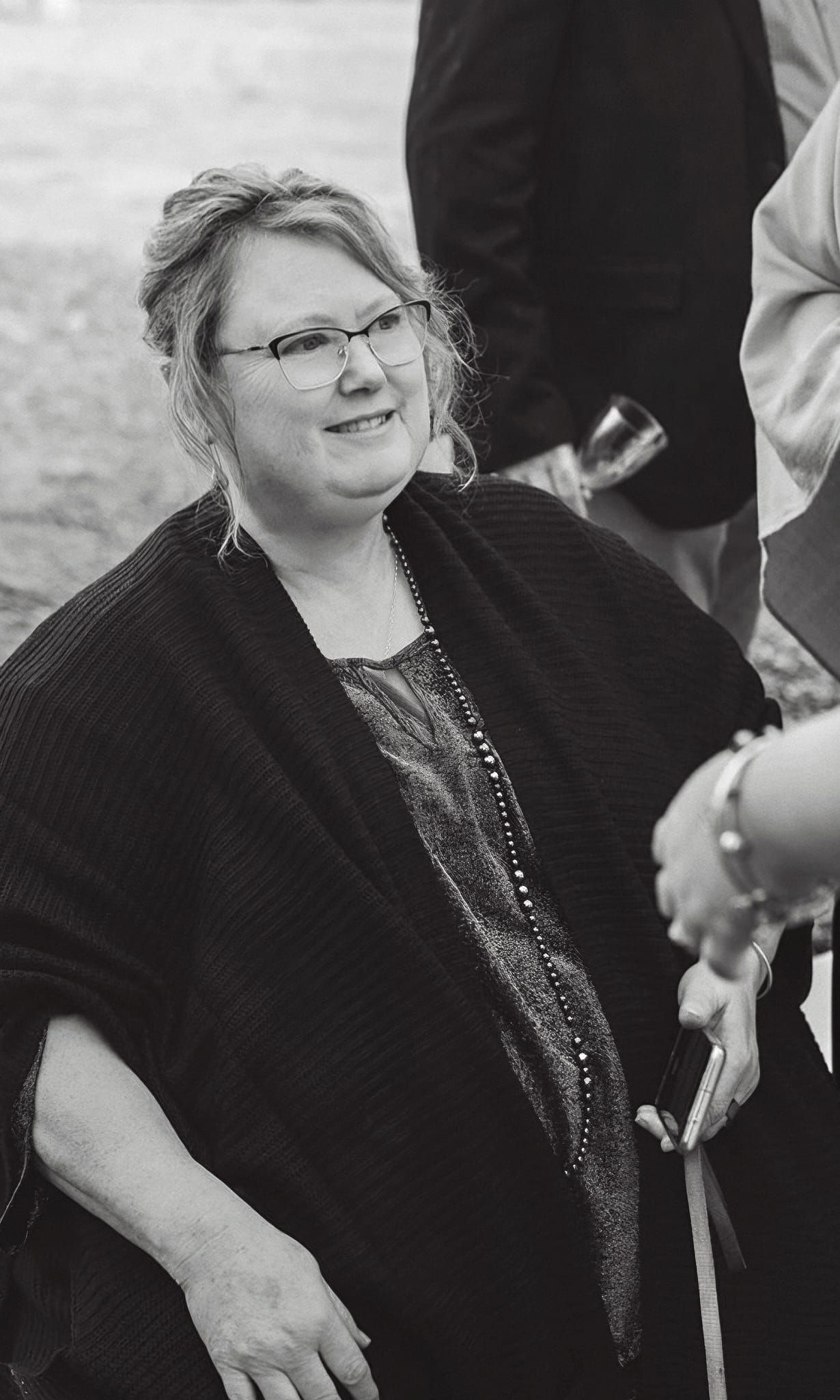 Smiling middle-aged woman with glasses and black shawl holding a cane at outdoor event.