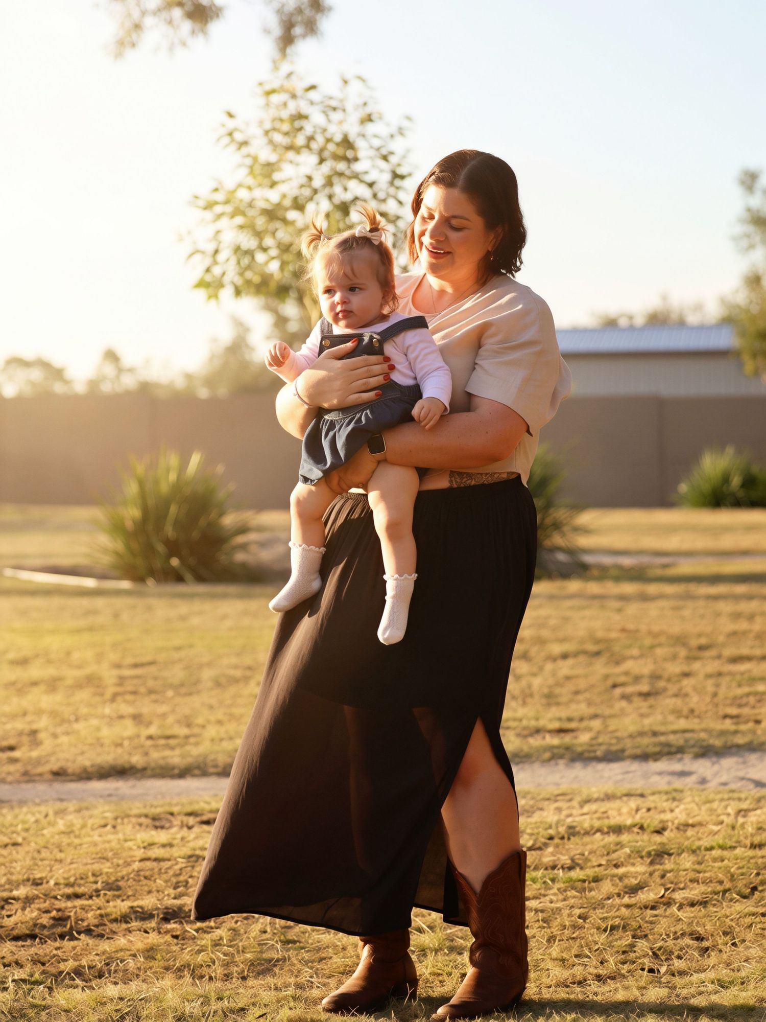 Mother holding baby girl in denim dress during golden hour outdoor family portrait.