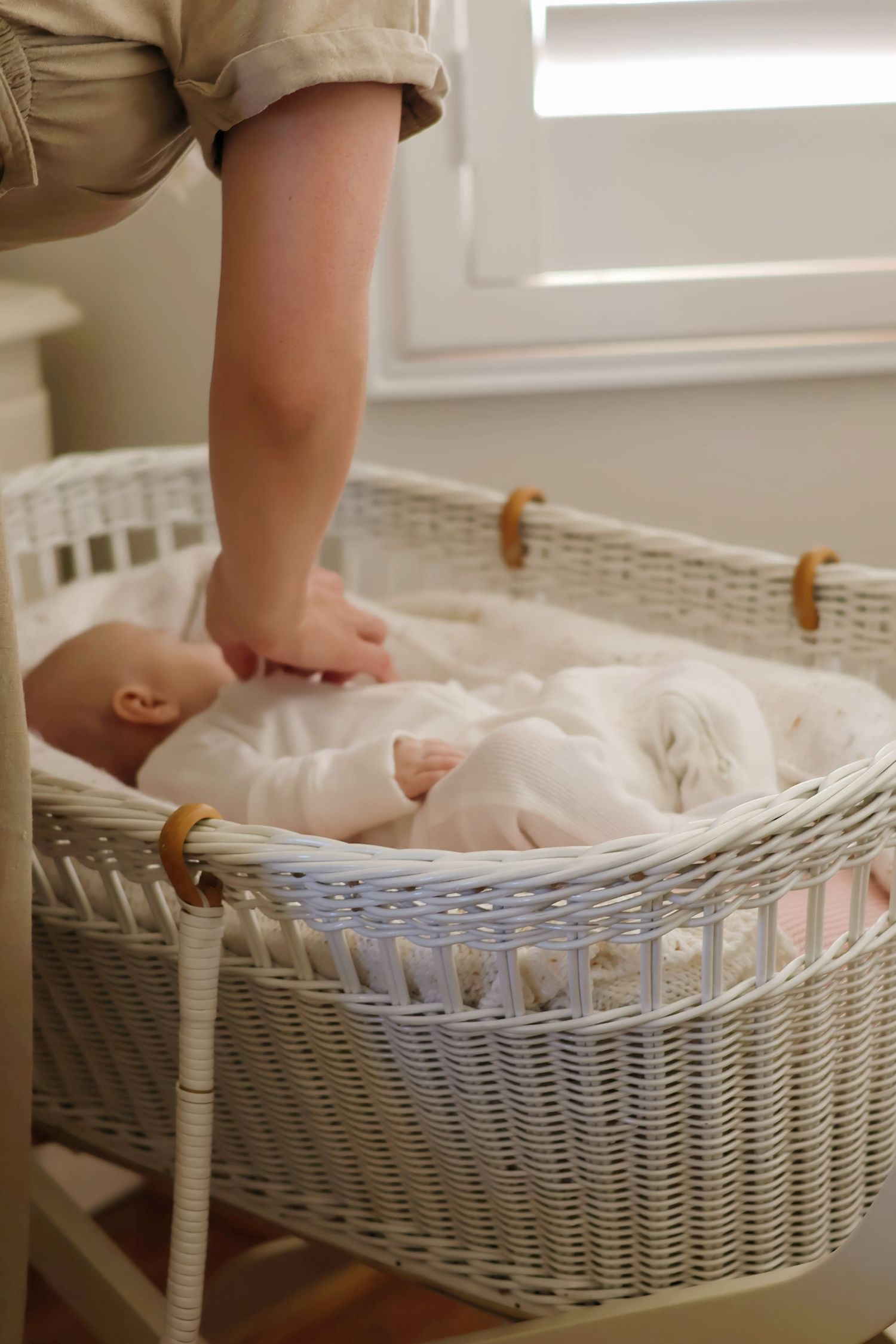 Newborn baby sleeping peacefully in a white wicker bassinet while a parent gently reaches in.