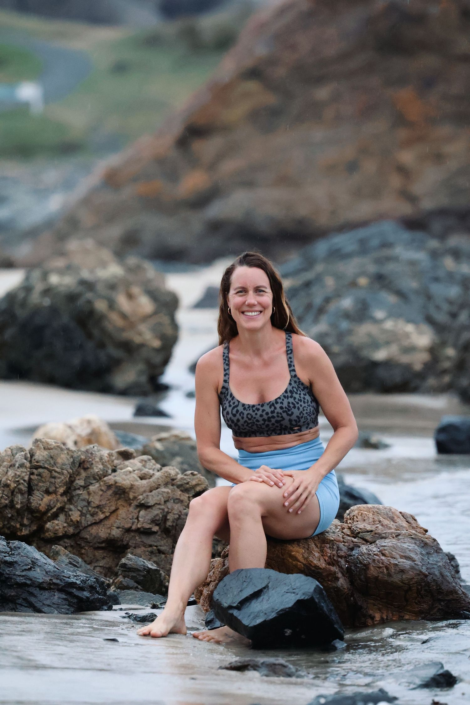 Athletic woman in sports bra and shorts sits barefoot on coastal rocks at the beach.