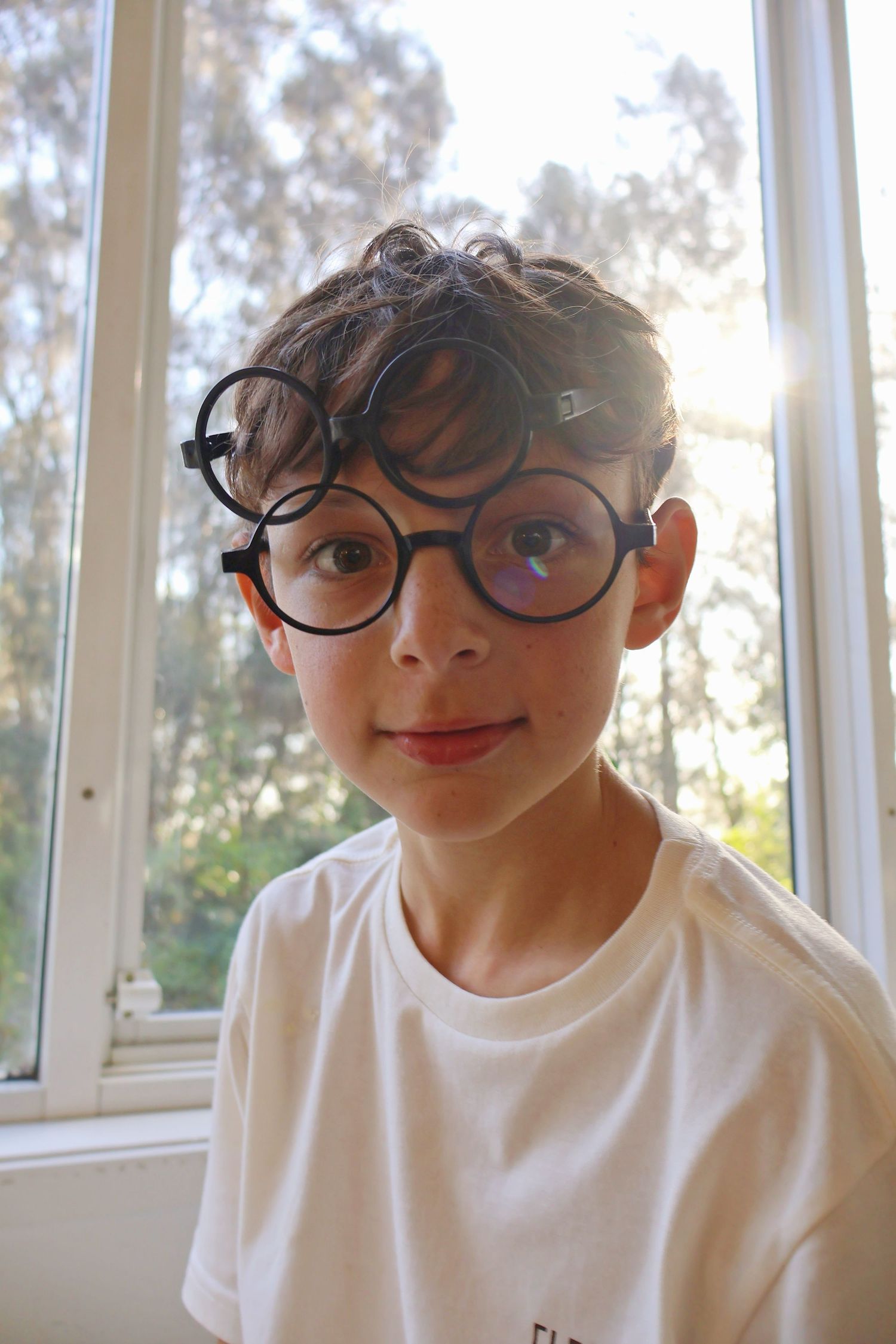 A young boy wearing oversized round Harry Potter glasses smiles in front of a bright window.