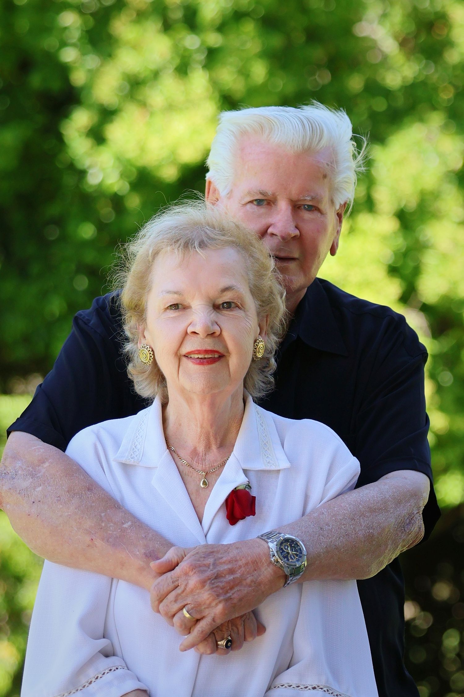 Happy senior couple embracing outdoors with lush green trees in background, man wearing watch and woman with red brooch.
