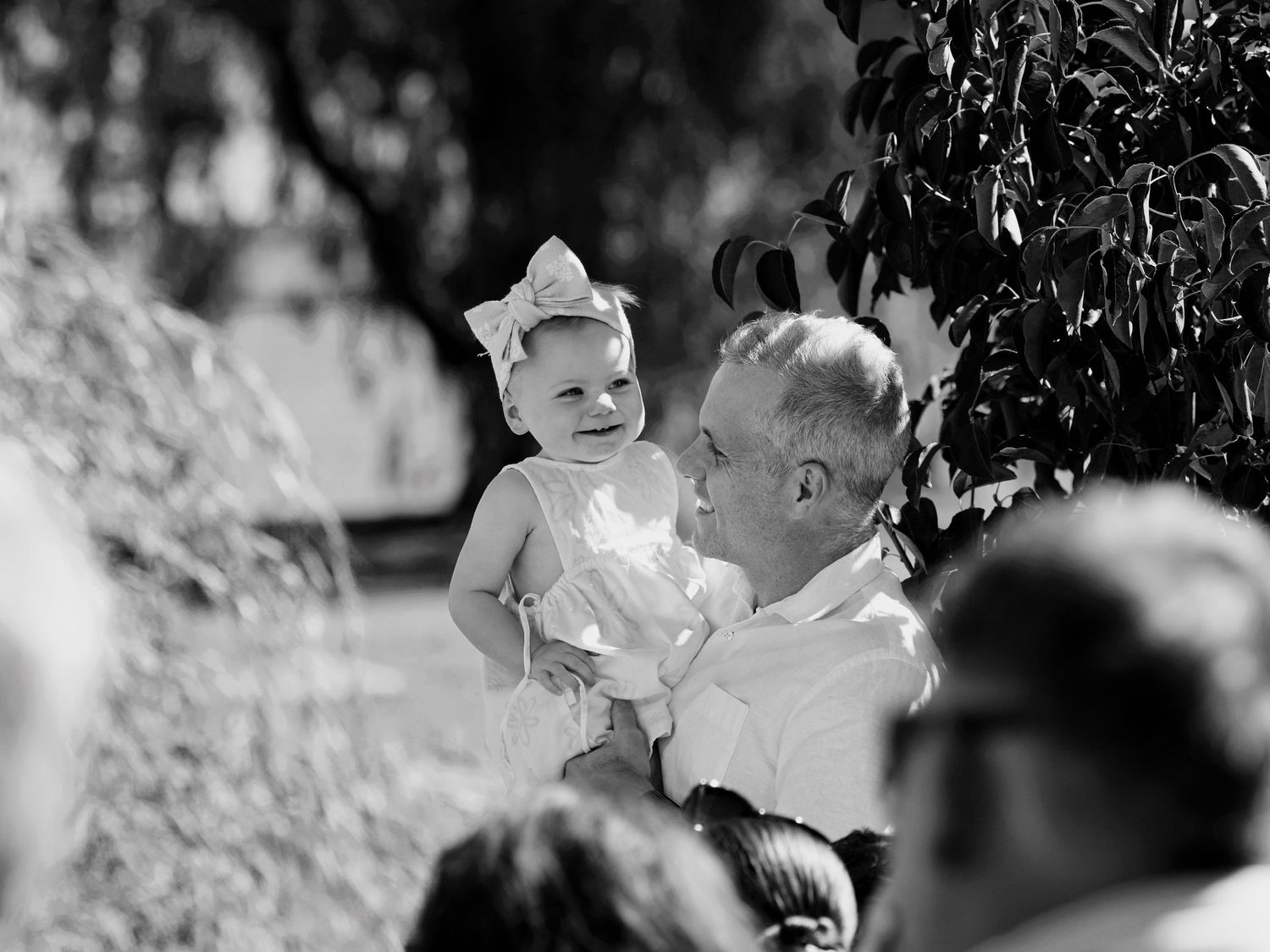 Black and white photo of a smiling grandfather holding a happy baby girl with a bow at an outdoor gathering.