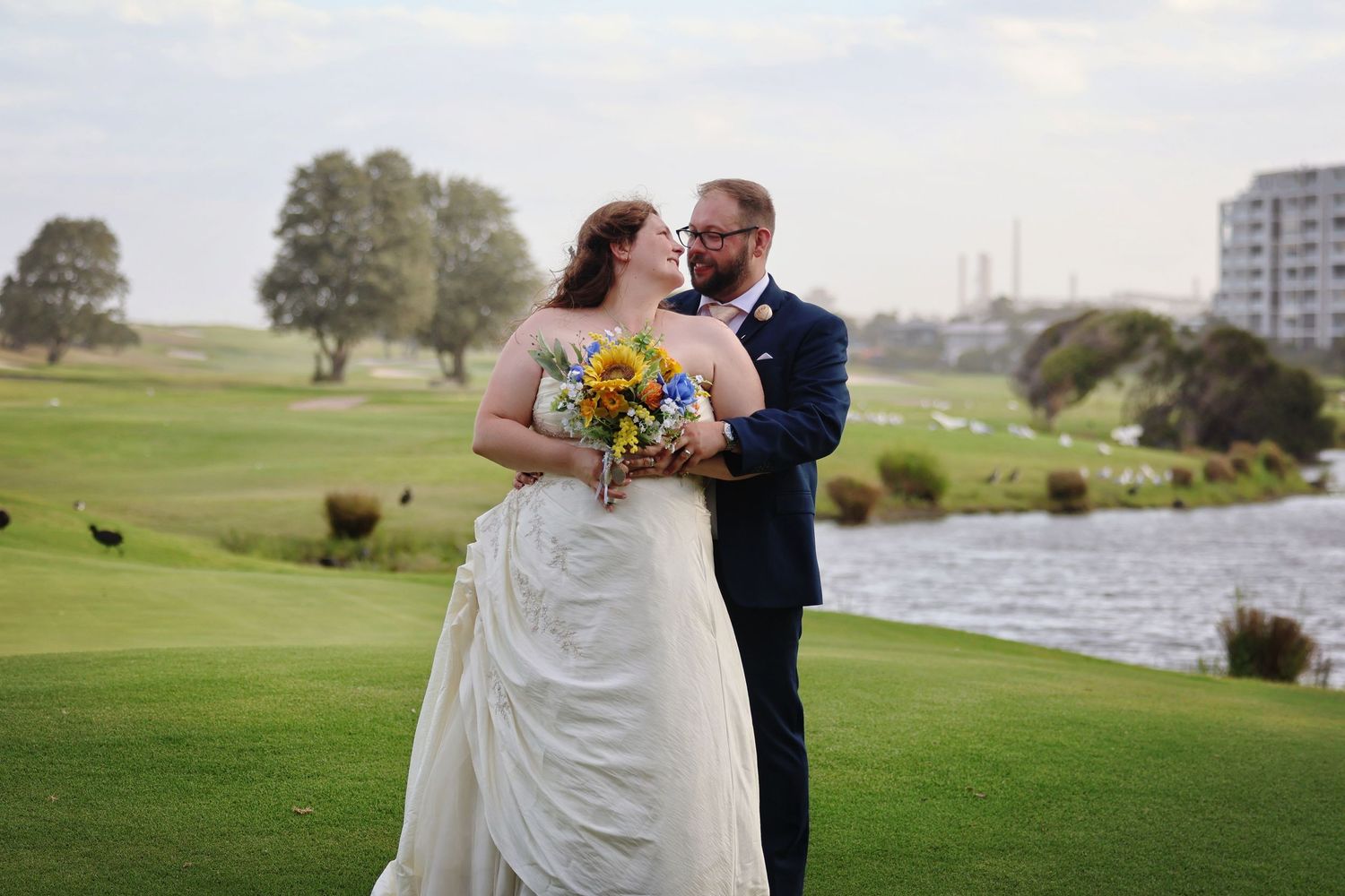 Bride and groom share romantic moment on golf course with scenic water backdrop.