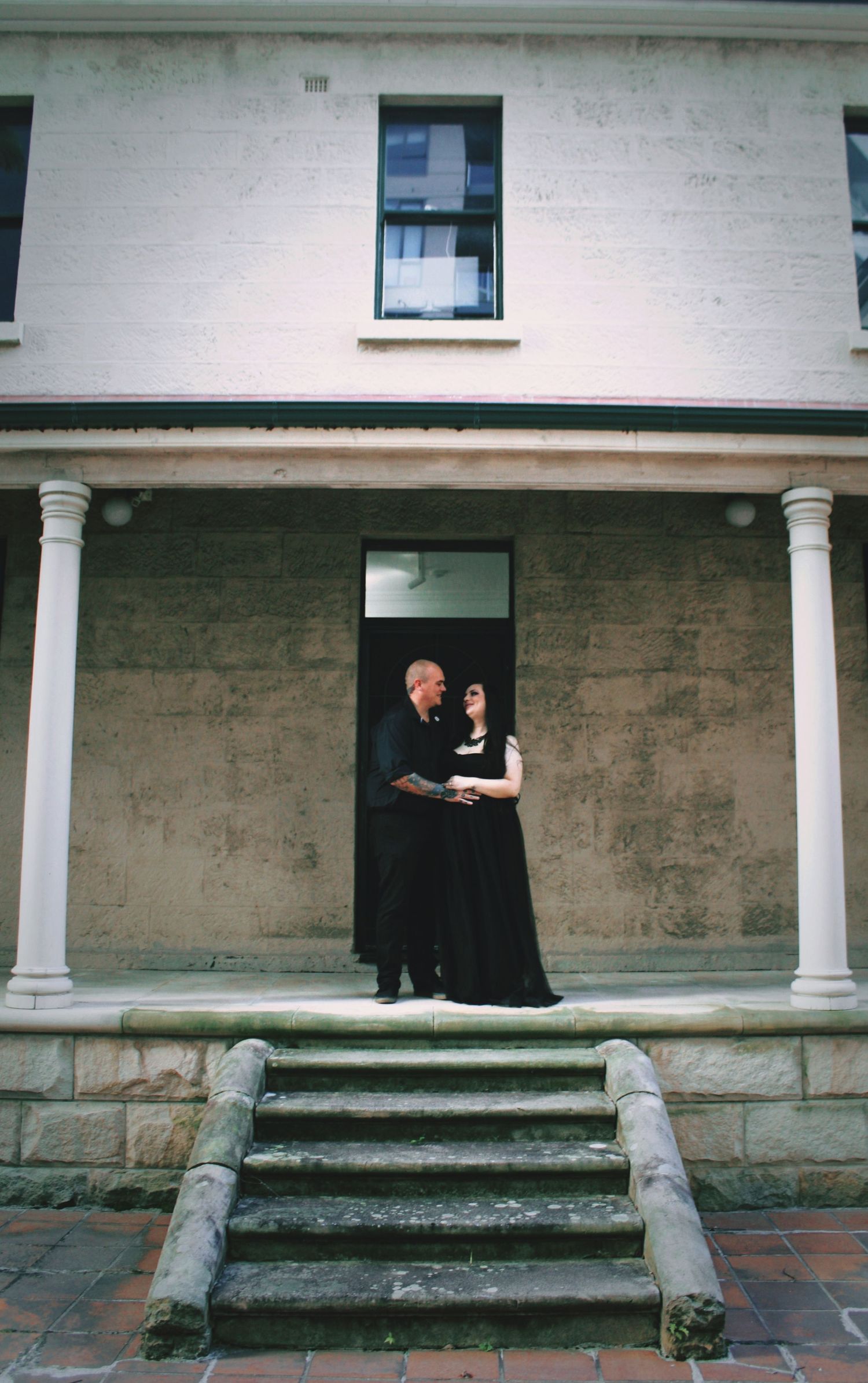 Couple in black wedding attire standing at entrance of historic white colonial building.