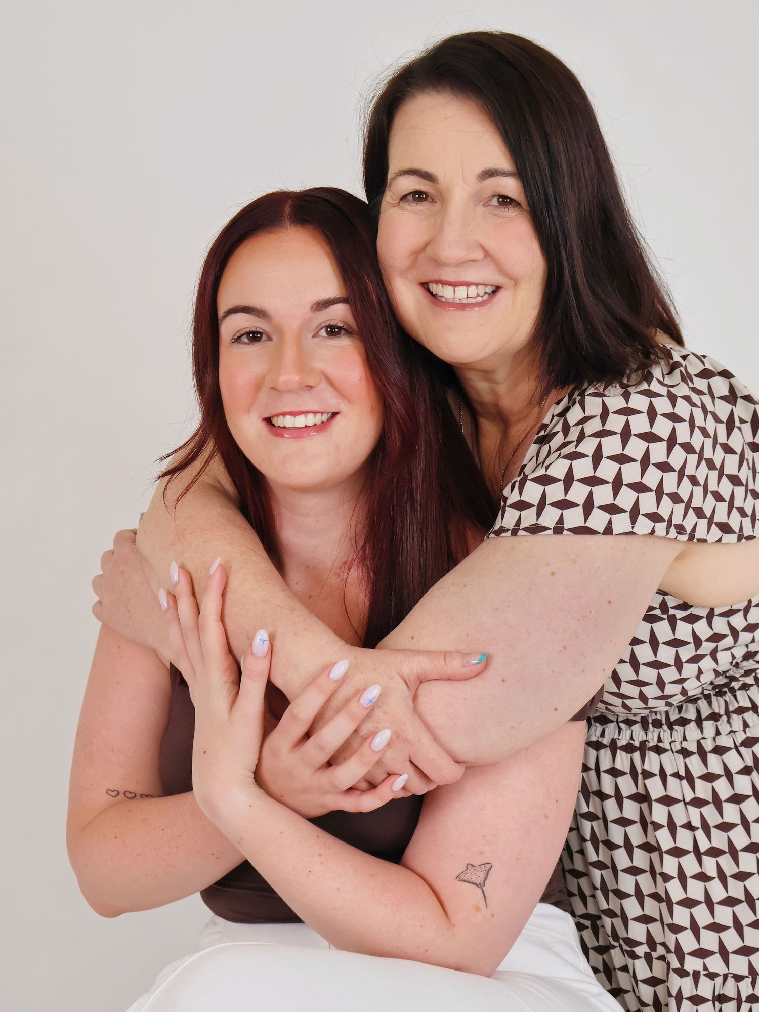 Two women smiling and embracing in a studio portrait, one with red hair and the other in a patterned top.