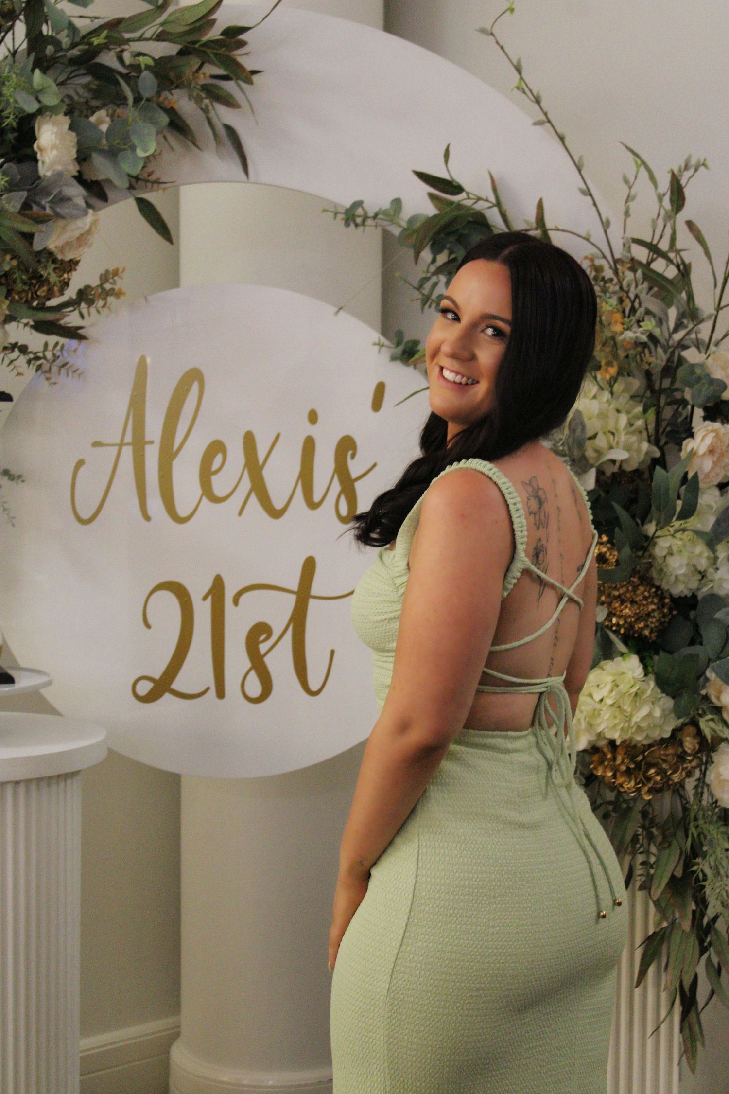 Young woman in green dress posing at Alexis 21st birthday party floral backdrop.