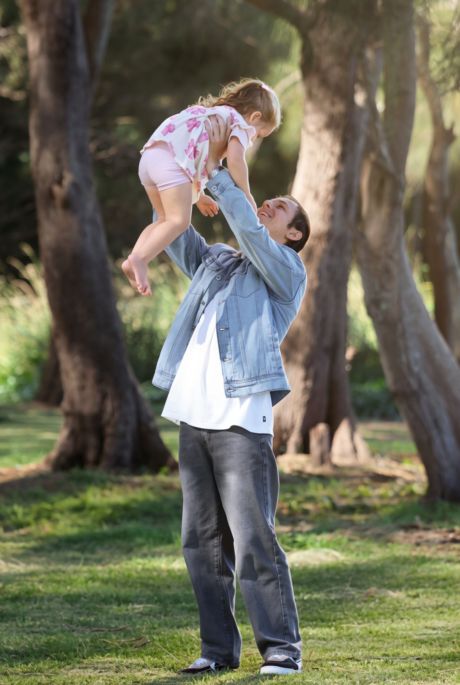 A father lifts his young daughter high in the air in a sunlit park surrounded by tall trees.