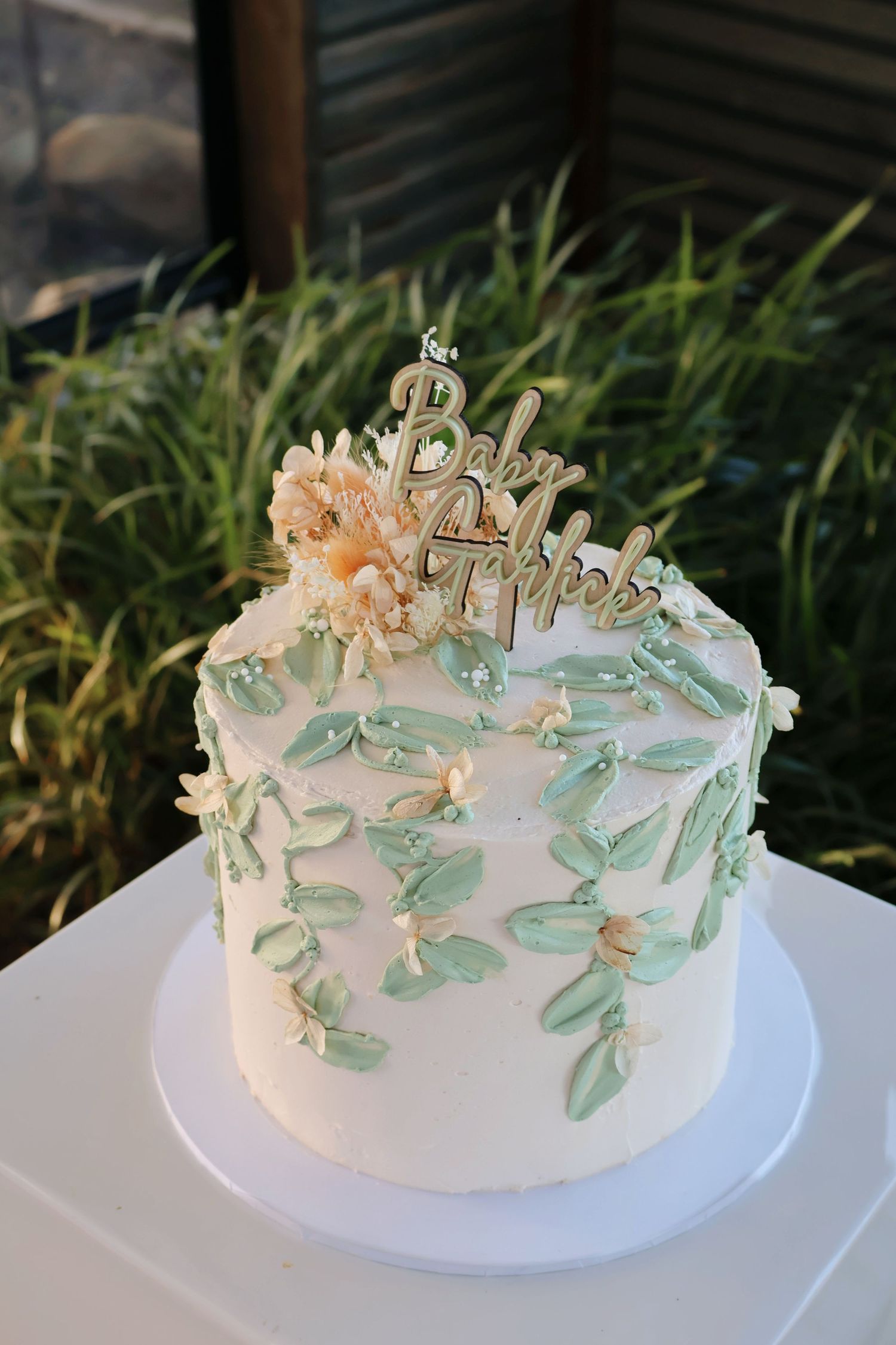 White baby shower cake with mint green leaf details and floral topper outdoors.