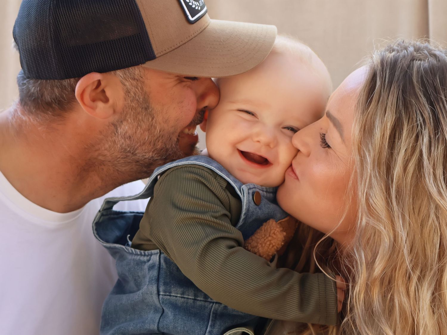 Happy parents kissing their laughing baby on the cheeks in a joyful family portrait outdoors.
