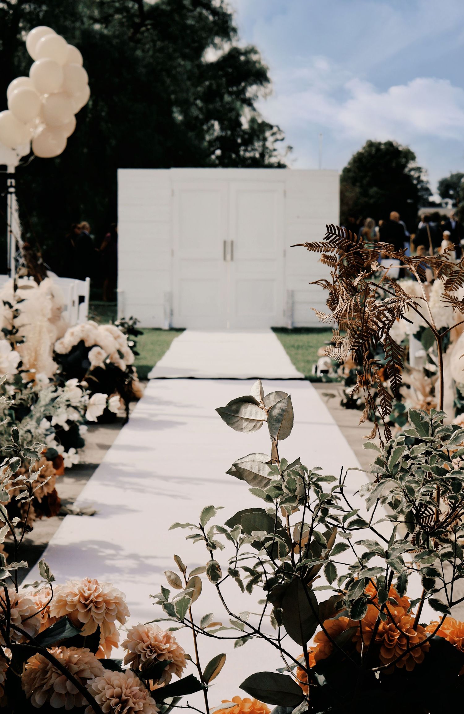 Elegant outdoor wedding aisle lined with white and amber florals leading to white doors backdrop under blue sky.