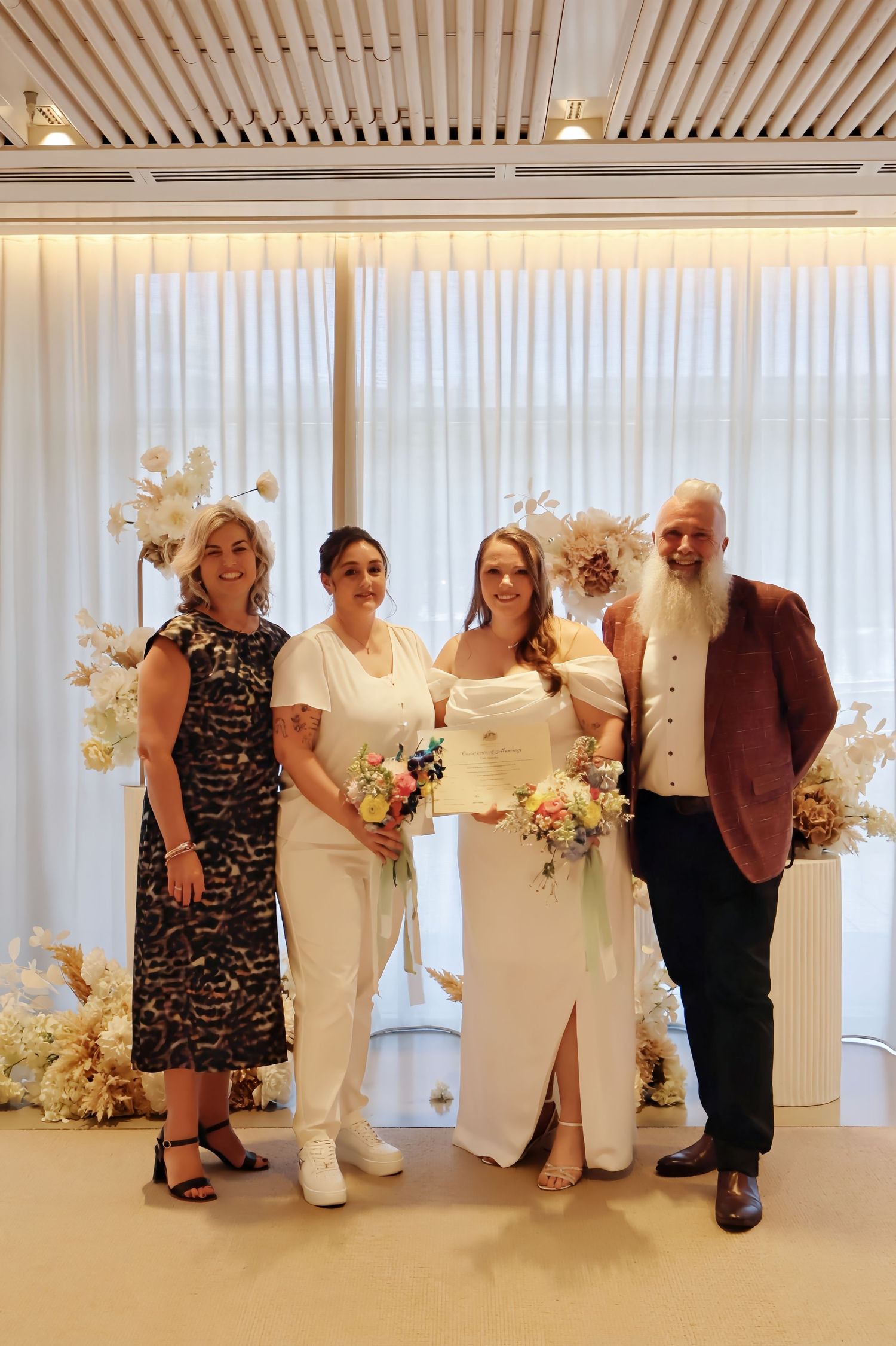 Two brides with bouquets posing with guests at modern wedding venue with white floral decor.
