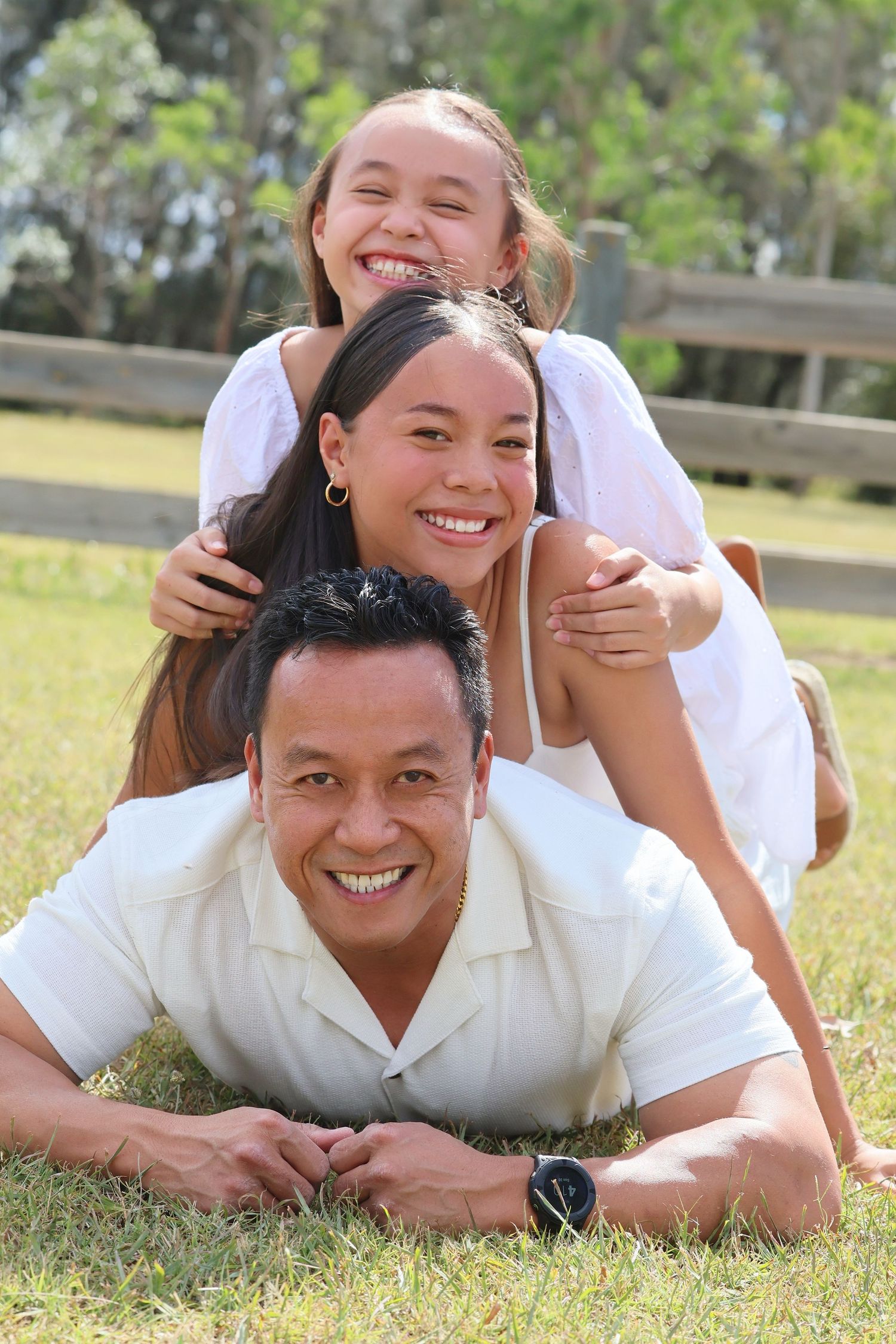 Smiling family of three stacking playfully on grass in a sunny outdoor park setting.