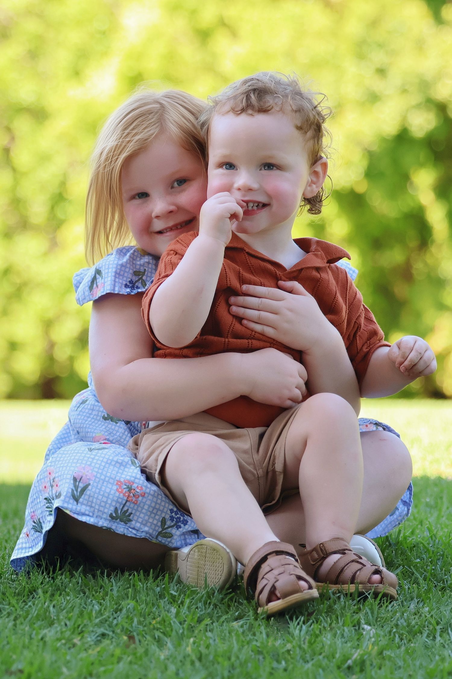 Two young siblings hugging and smiling while sitting together on green grass outdoors.