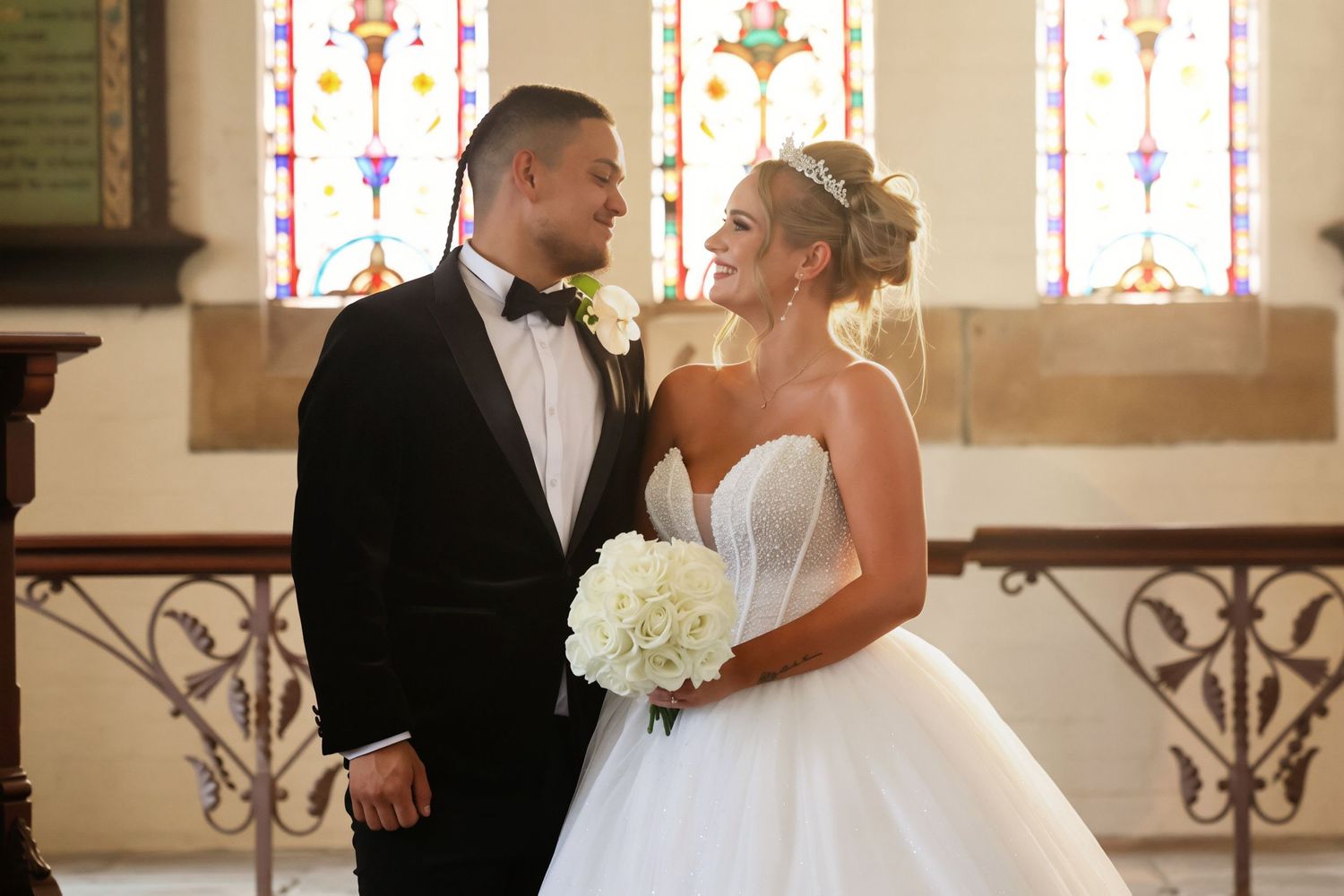 Bride and groom share a loving glance inside a church, bride holding white rose bouquet.
