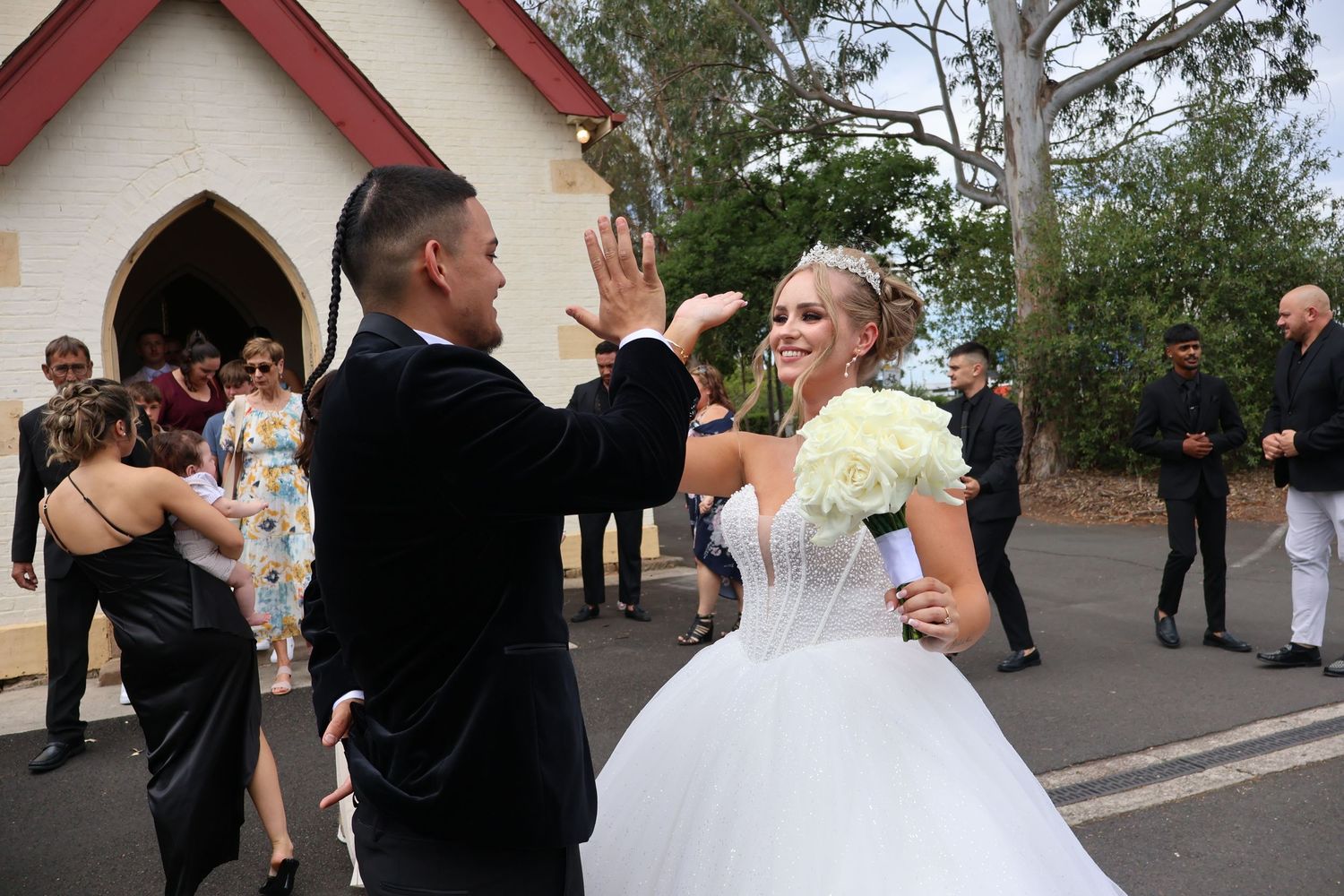 Bride in white ball gown and groom celebrate outside a chapel, guests applauding in background.