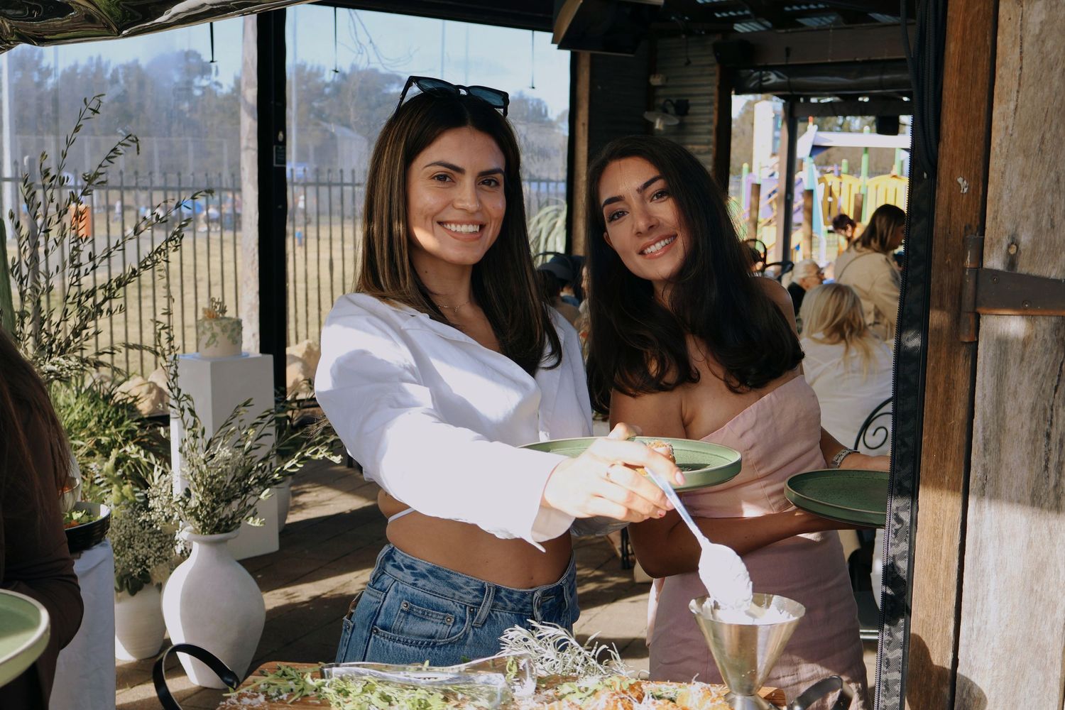 Two smiling women serving food at an outdoor baby shower grazing table.