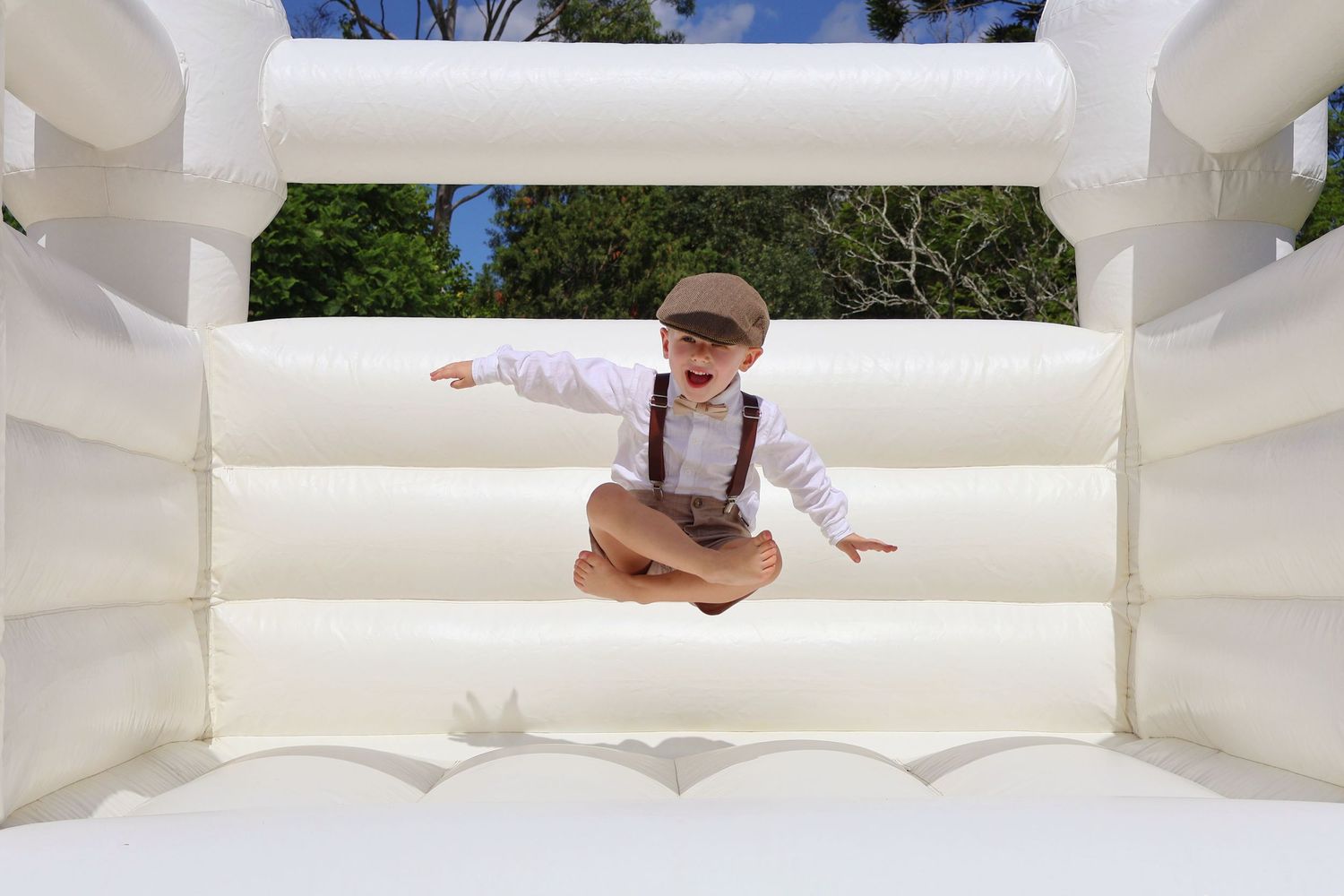 Young boy in suspenders jumping joyfully in white inflatable bounce house outdoors.