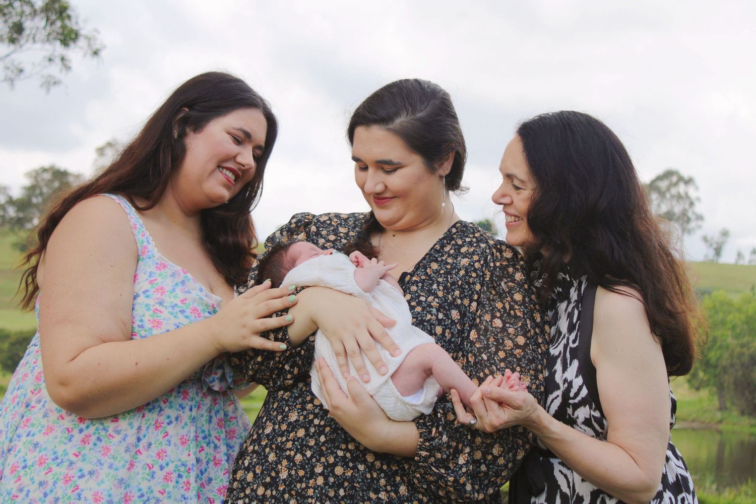 Three women admire a newborn baby outdoors near a lush green park setting.