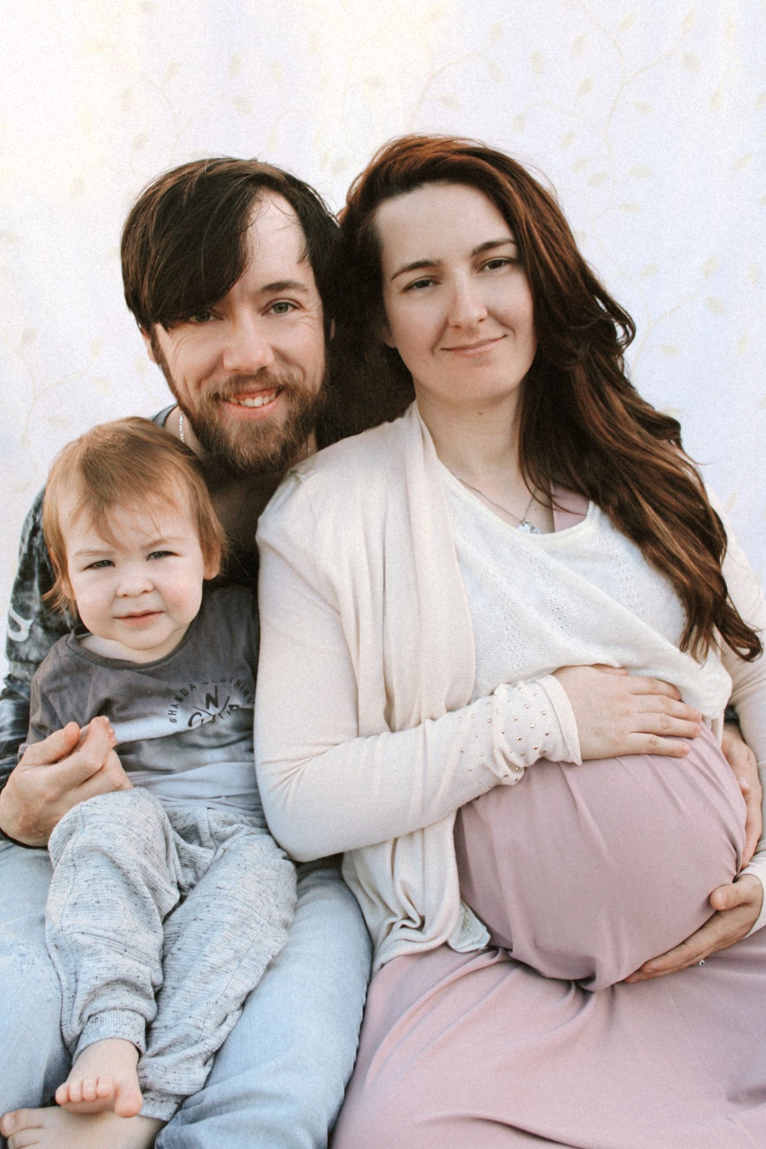 Happy family maternity photo with father, toddler, and pregnant mother sitting together against white background.
