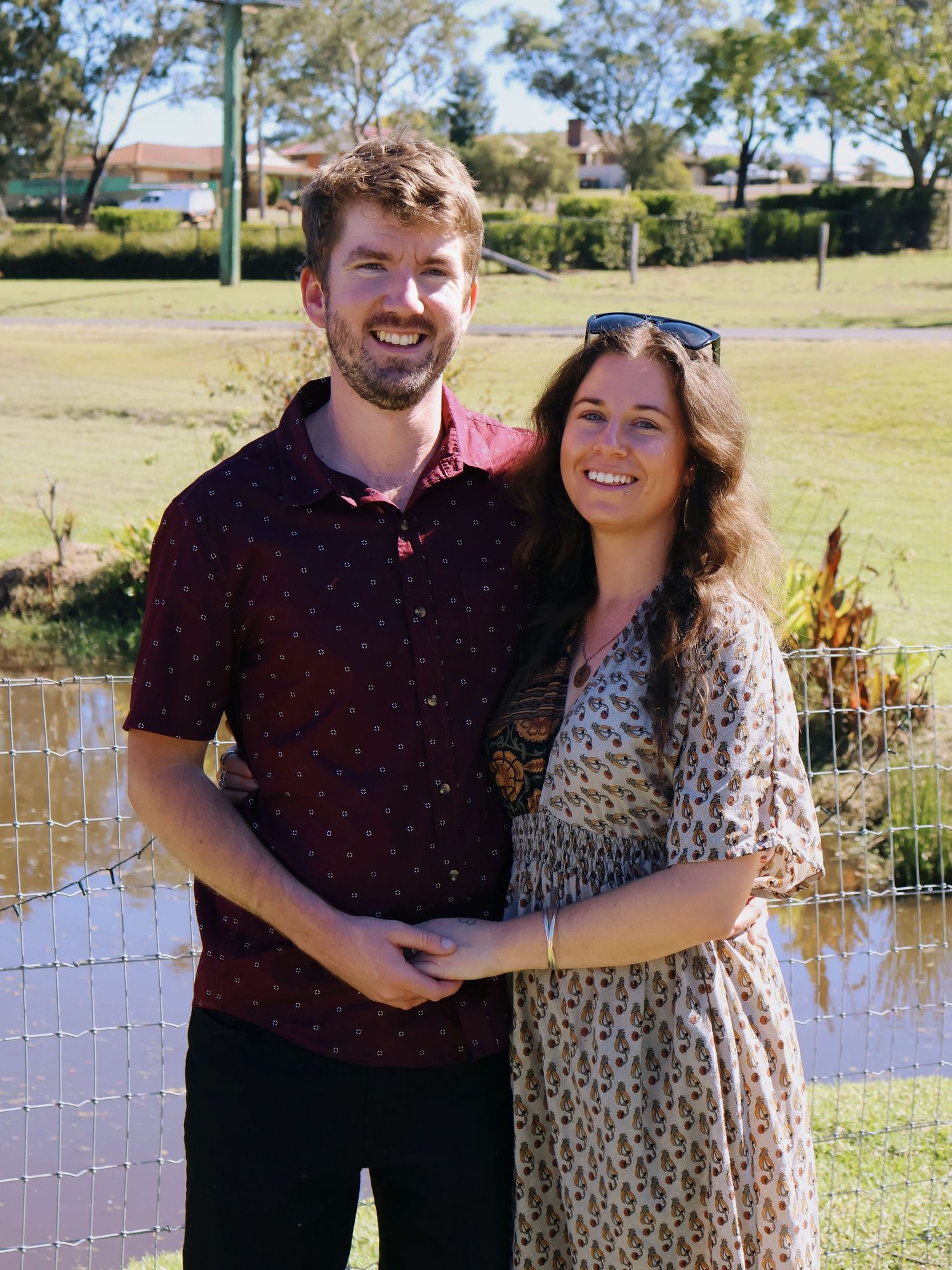 Smiling couple posing together outdoors near a pond in a sunny suburban backyard setting.