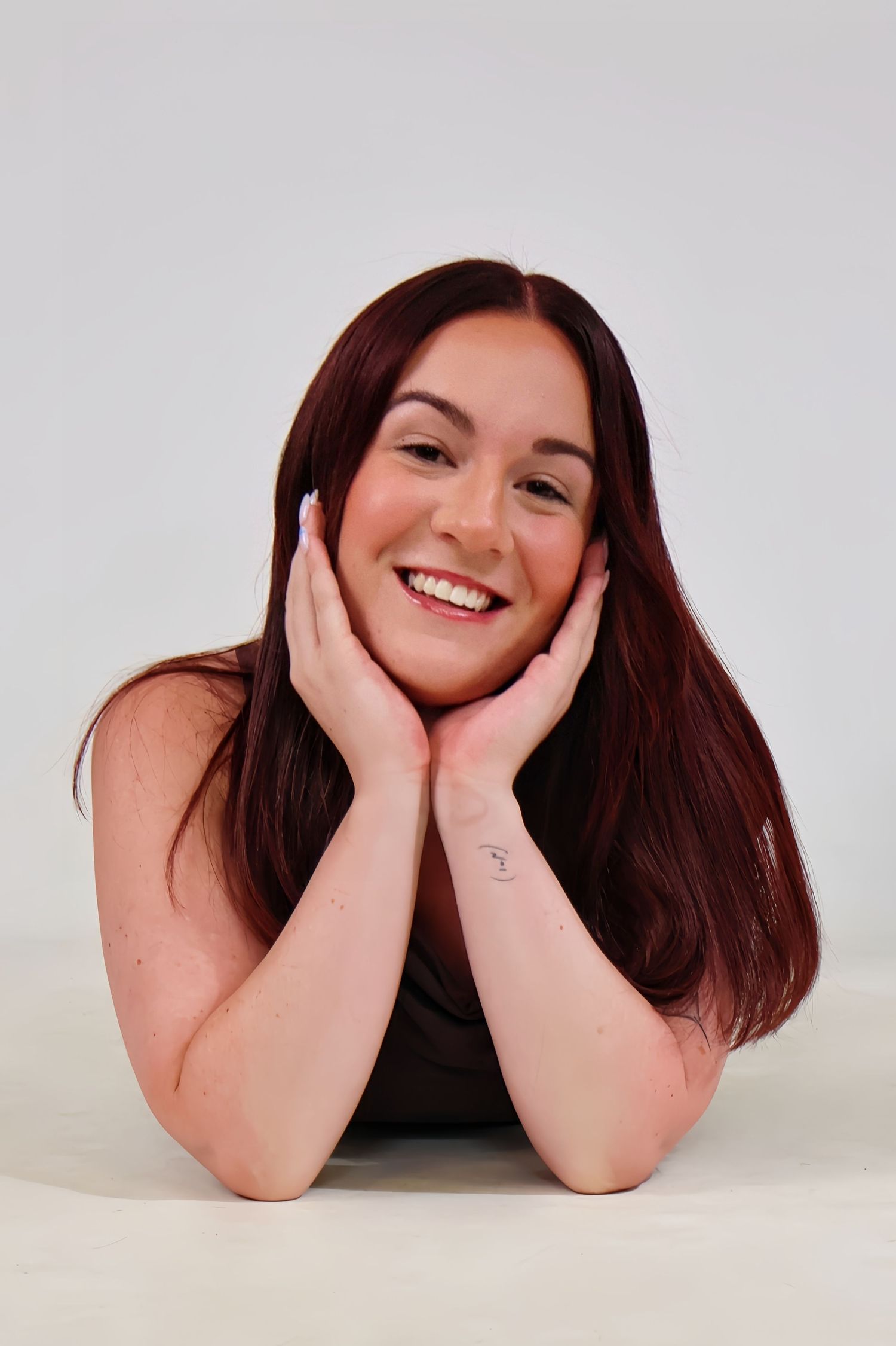 Young woman with long auburn hair smiling with hands on cheeks in white studio portrait.