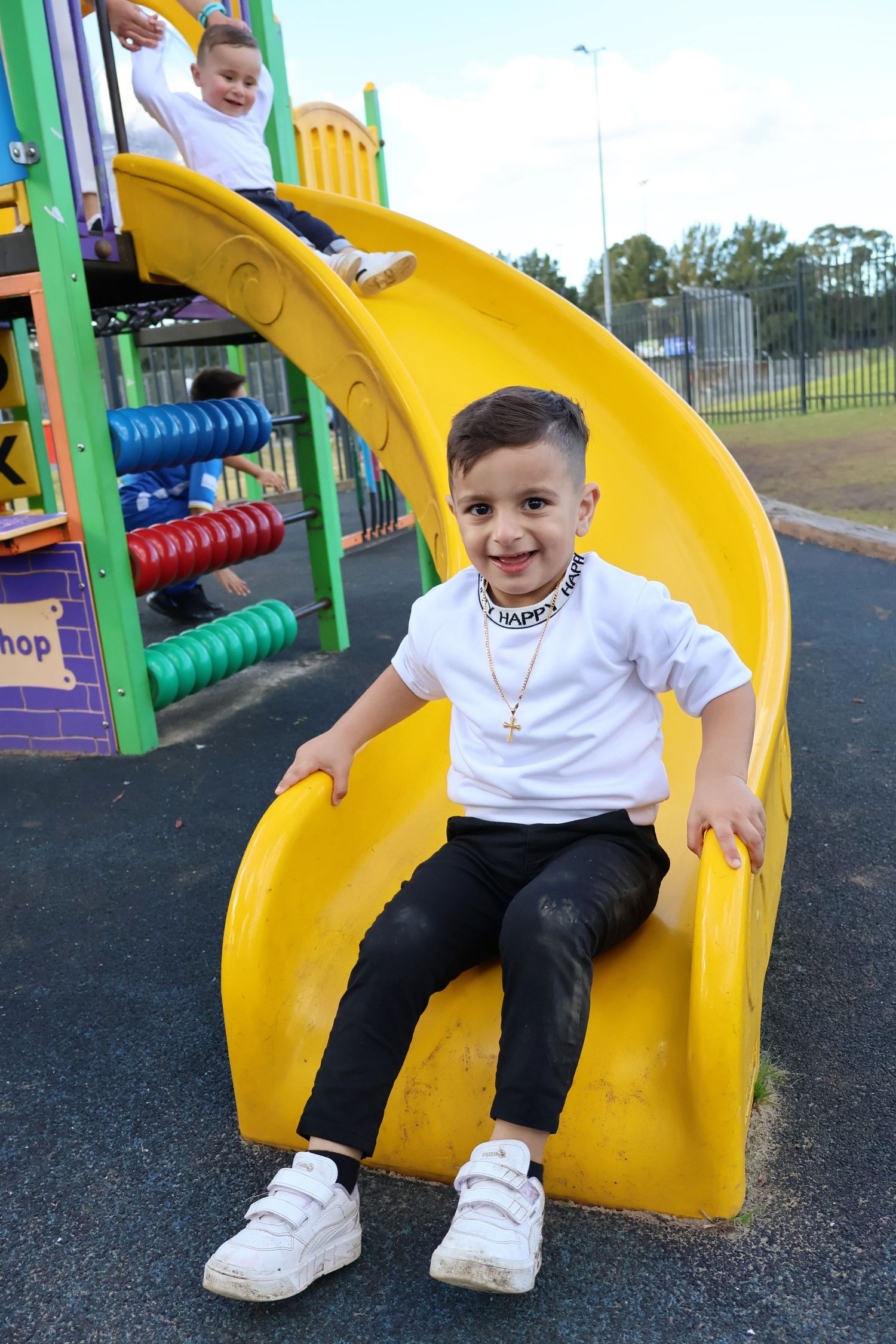Smiling young boy sitting on yellow playground slide wearing white shirt and necklace.