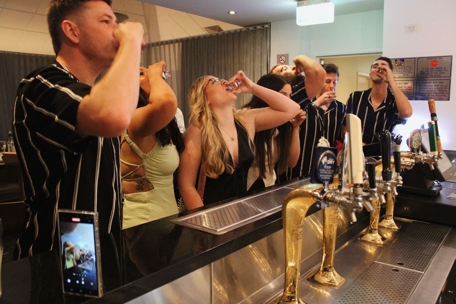 Bar staff doing shots together behind a row of gold beer taps at a modern bar.
