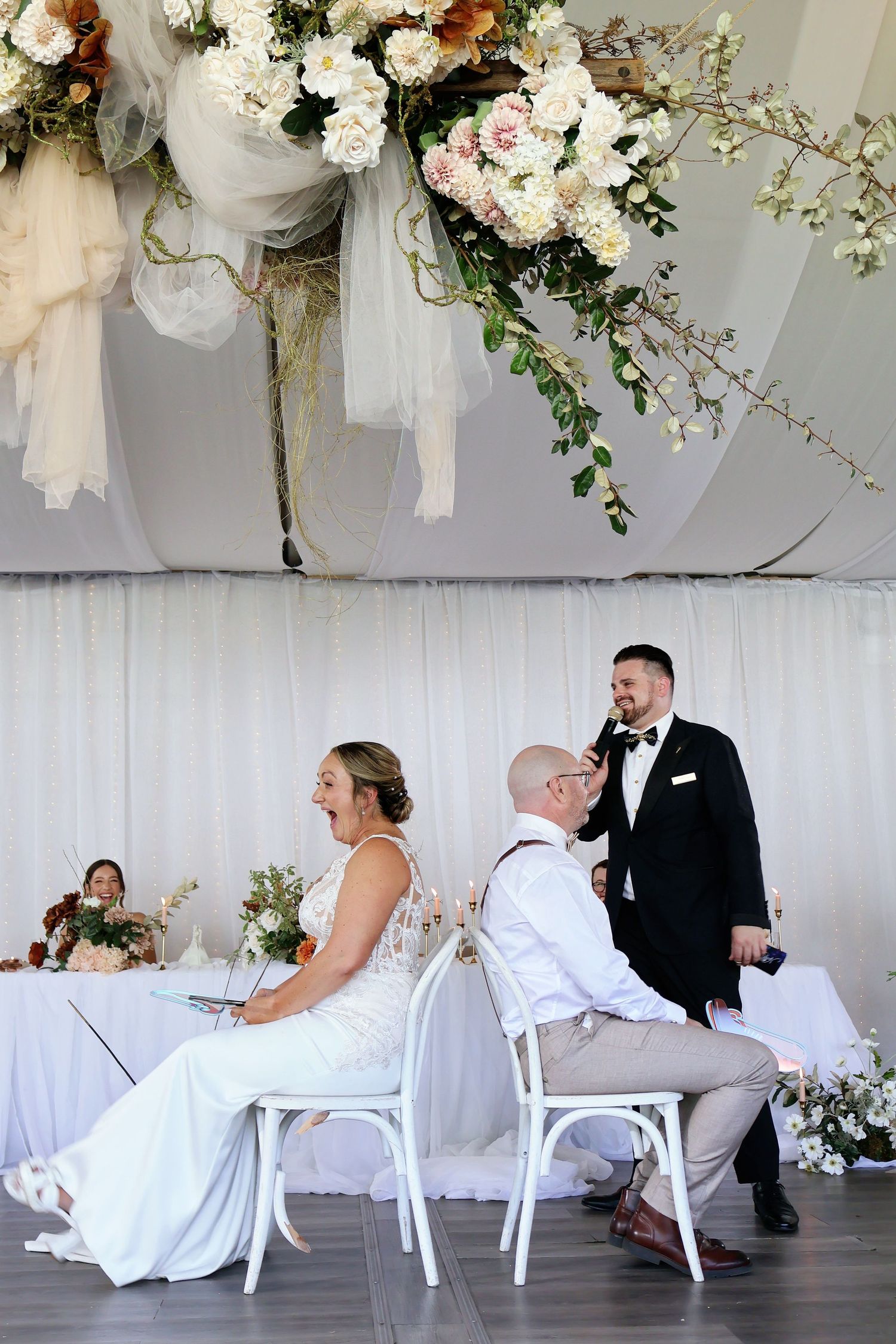 Bride laughing joyfully at reception table under elegant floral ceiling decorations.