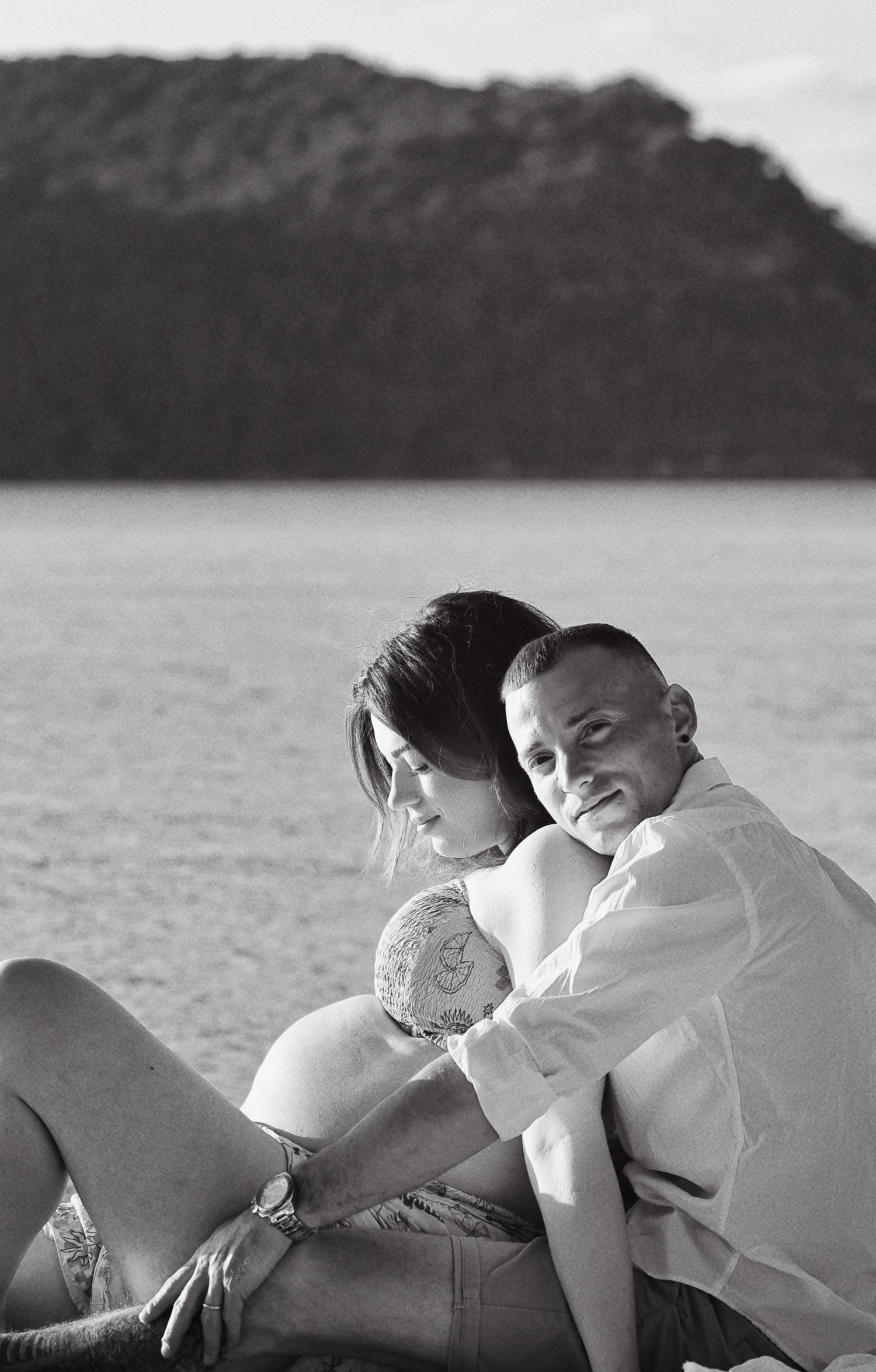 Black and white maternity photo of a couple sitting together on a beach by the water.