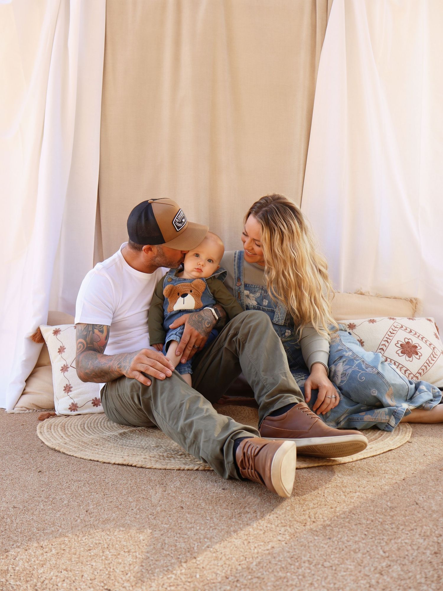 Young family with tattooed father, blonde mother, and baby boy sitting together in a boho tent setup.